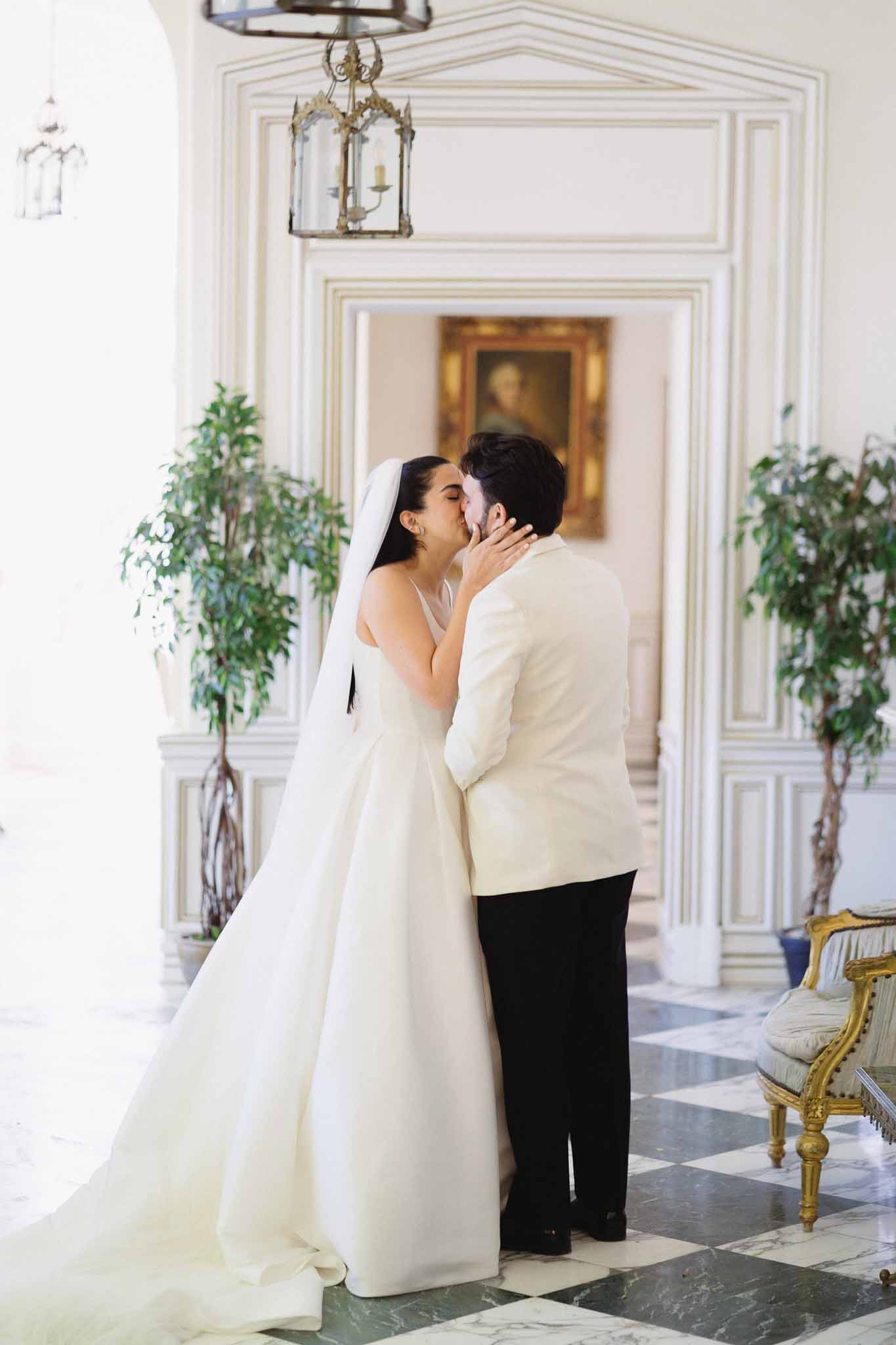 Couple kissing on marble checkerboard floor in boiserie hall with gilt portrait, lantern, and potted trees flanking doorway