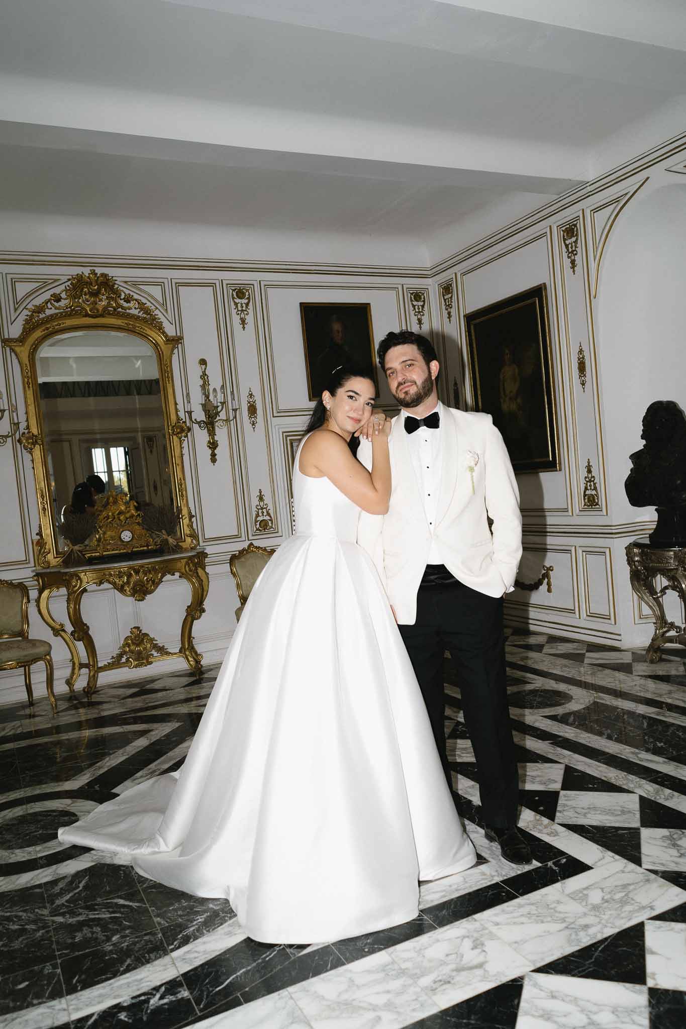 Bride in white satin ball gown leaning on groom in ivory dinner jacket in gilt-trimmed palace room