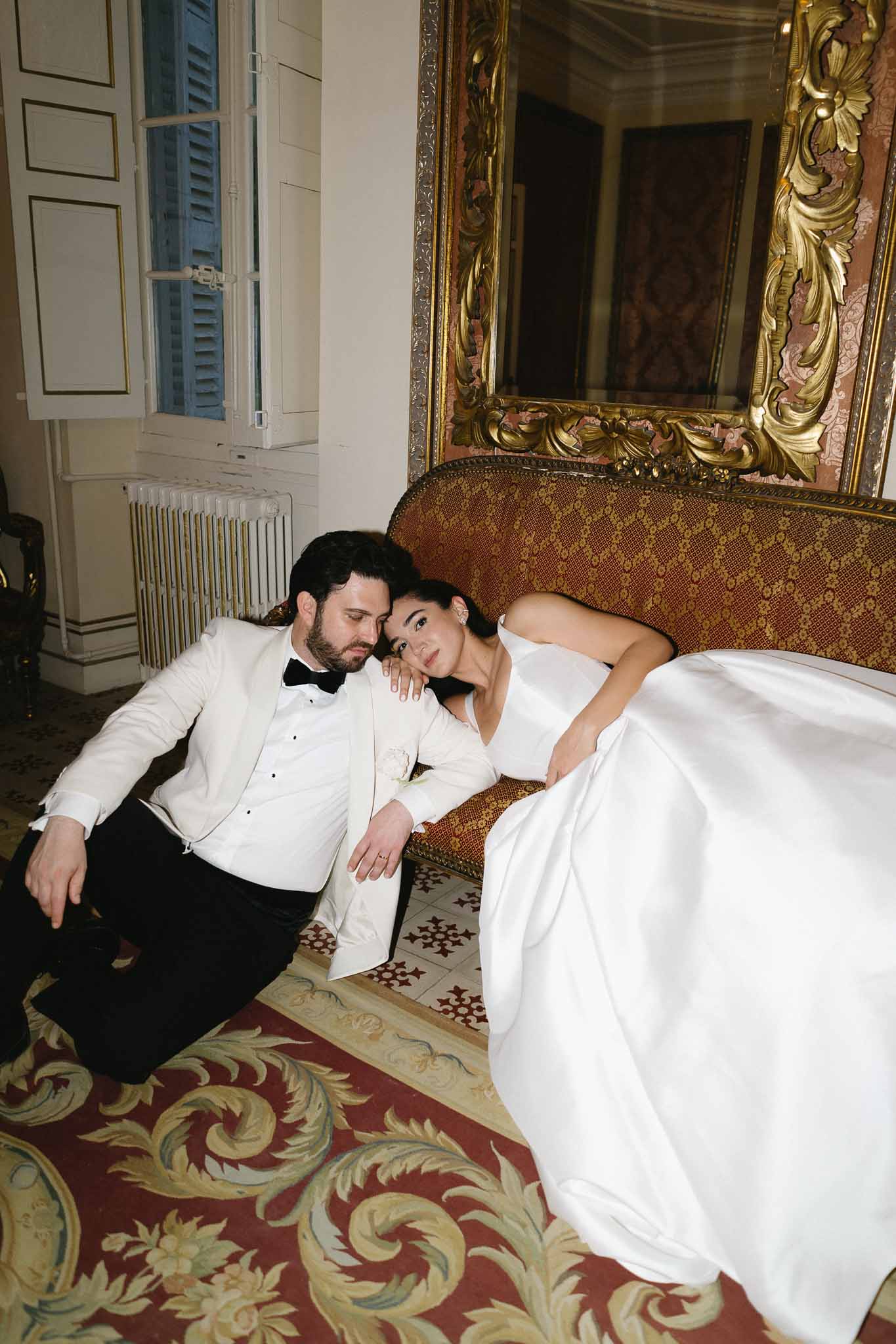 Groom on floor and bride reclining on antique red sofa before gilt baroque mirror in chateau salon