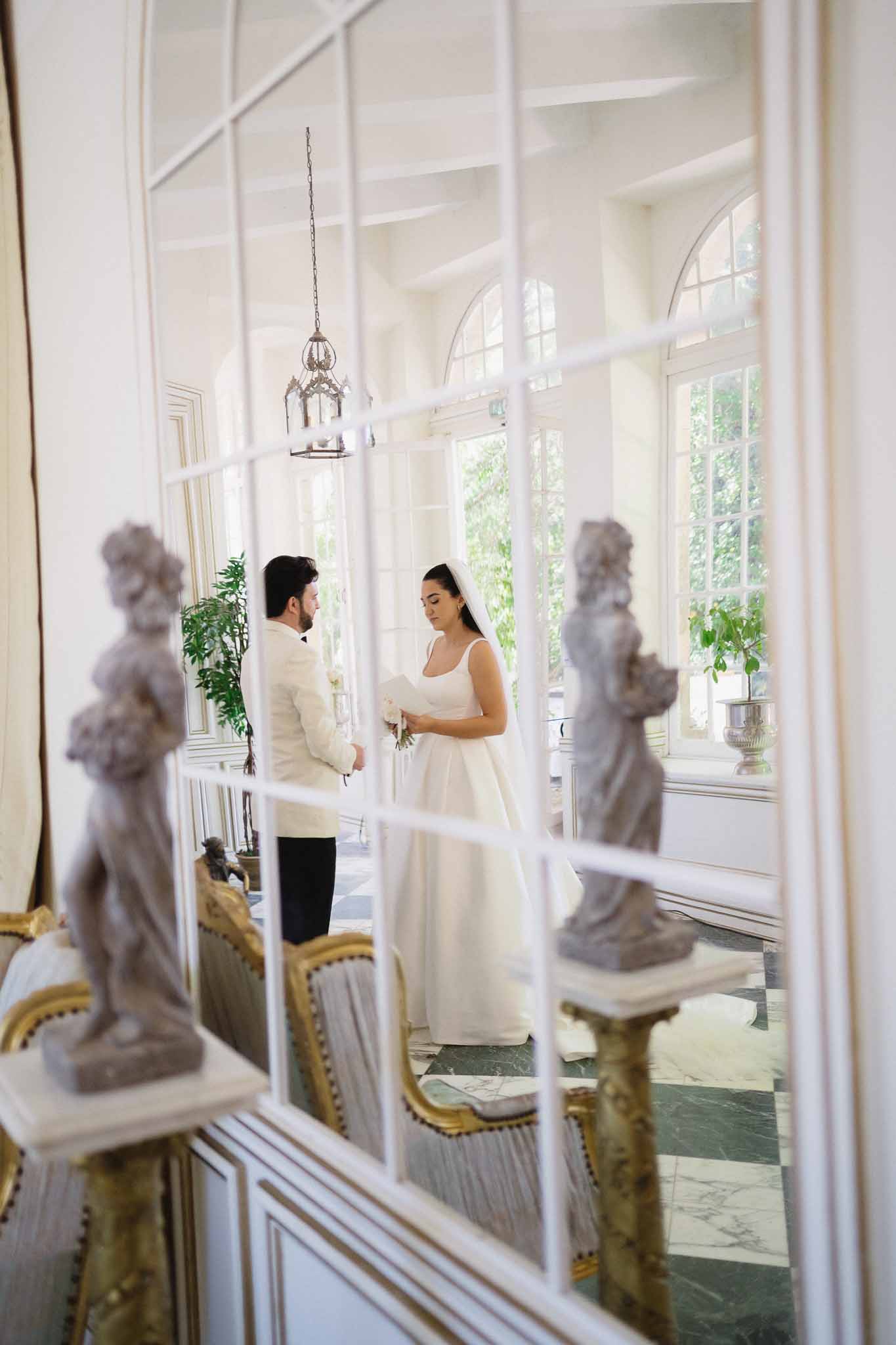 Couple exchanging vows in chateau interior with marble floor and stone sculptures shot through glass