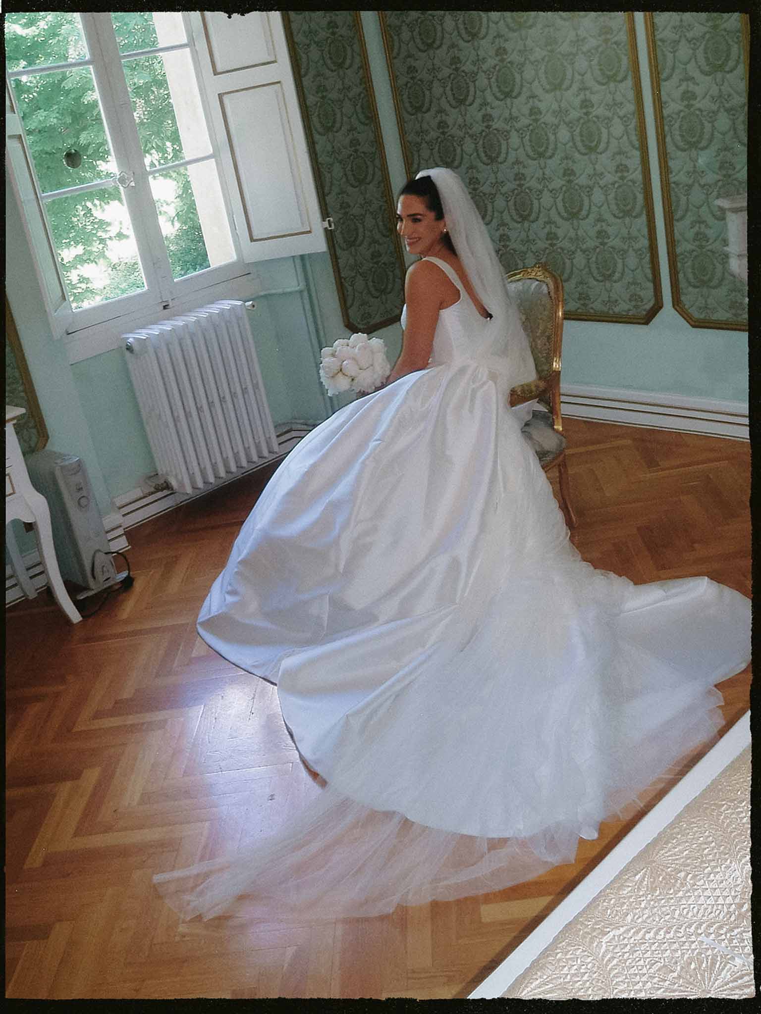 Bride with peony bouquet on gilt chair with cathedral train across parquet in blue-green paneled salon