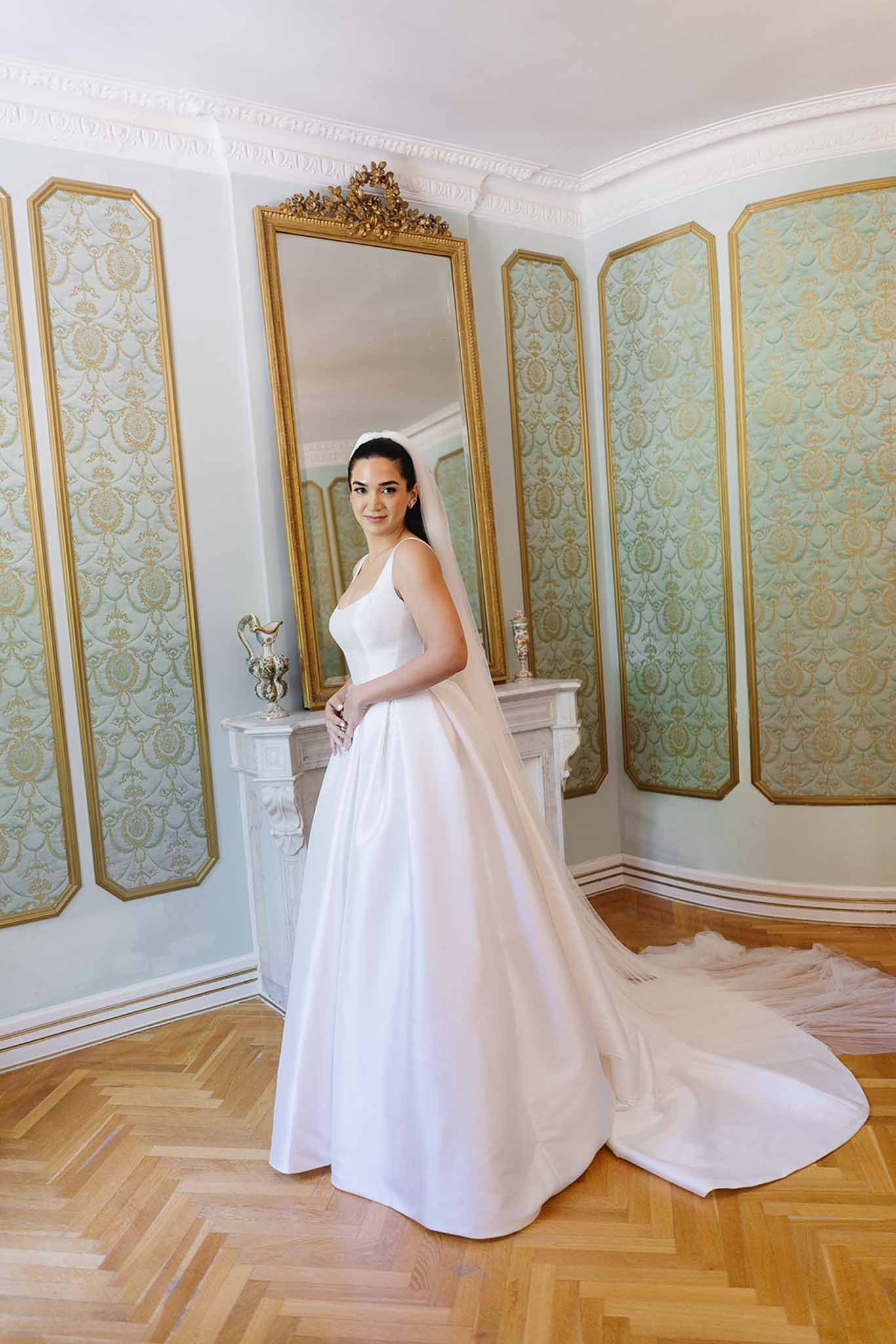 Bride in white satin ball gown with cathedral train and veil in ornate French room with gilt mirror