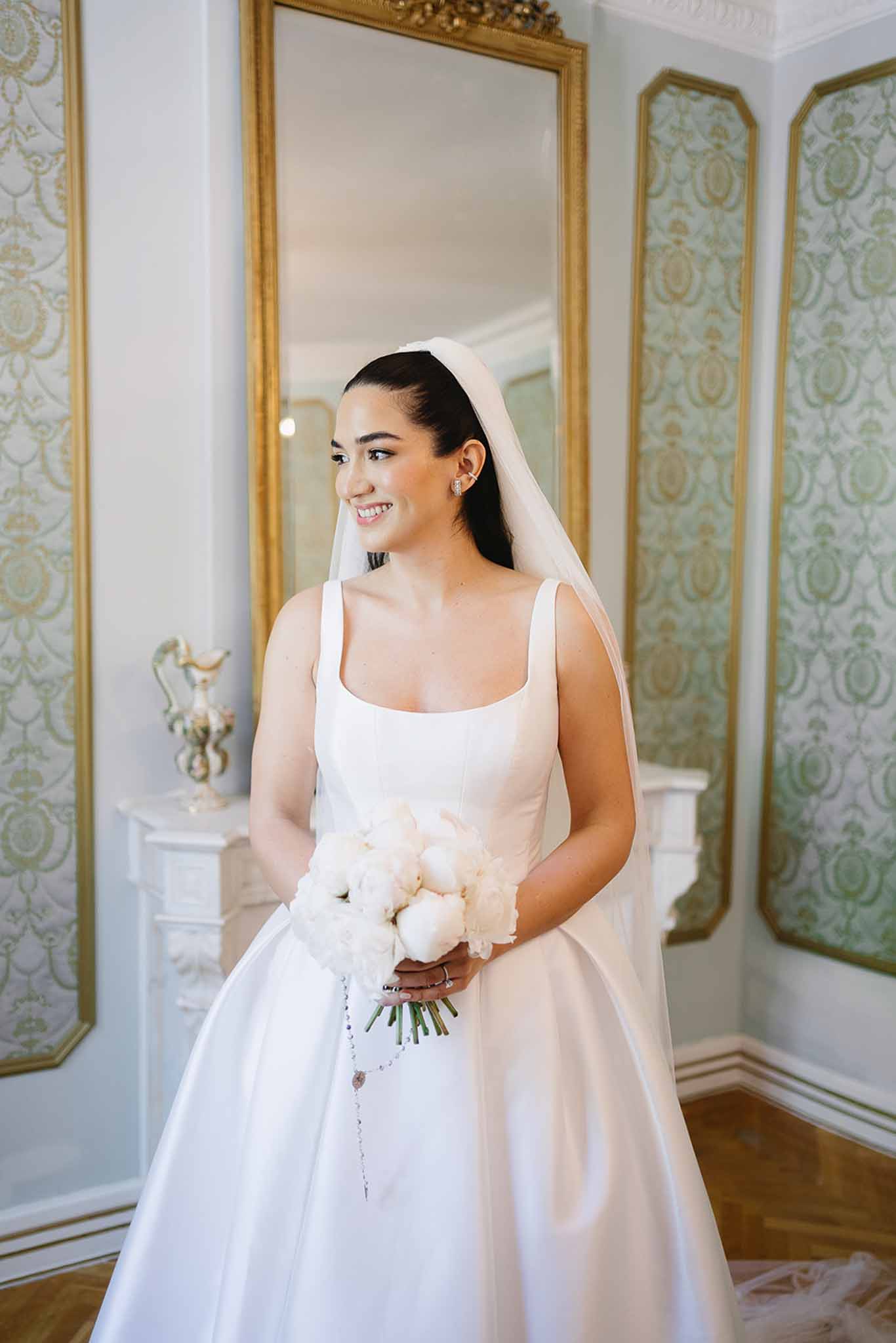 Bride in ivory satin ball gown with cathedral veil holding white peony bouquet in chateau room with gold mirrors
