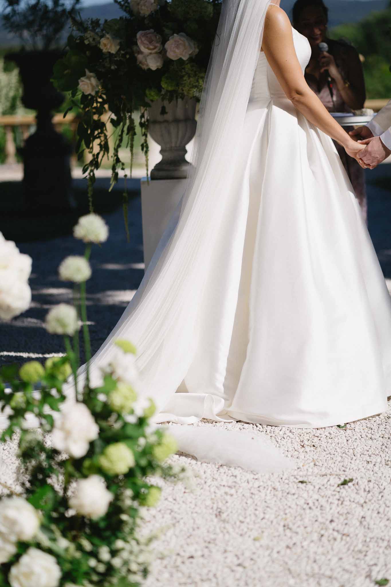 Waist-down detail of bride's satin gown and tulle veil with white ranunculus aisle arrangements