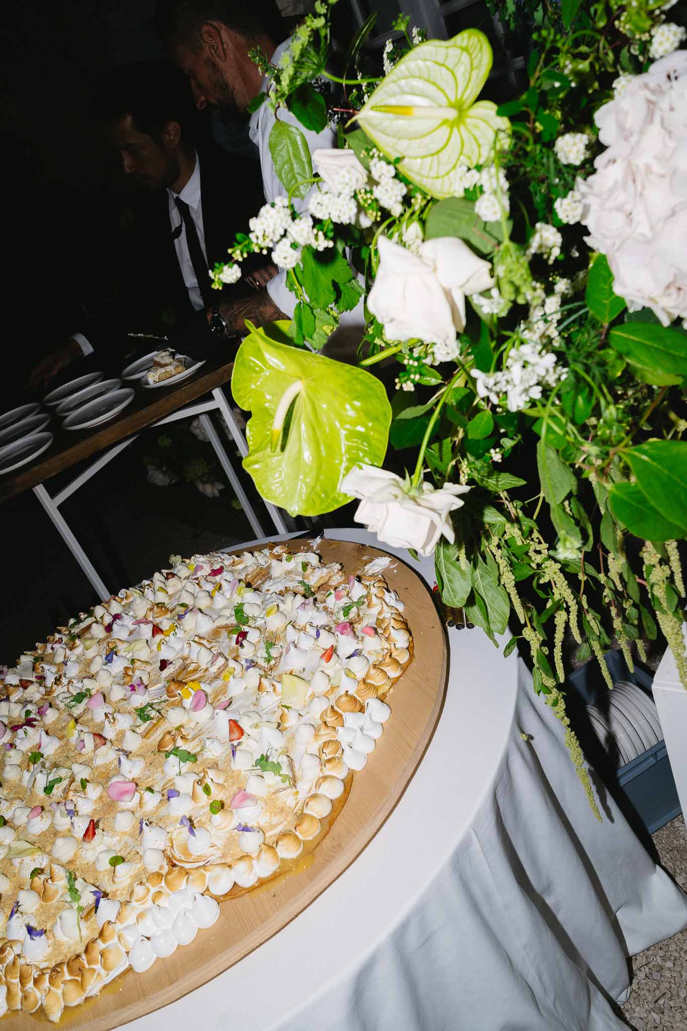 Oval meringue dessert with edible flowers beside white peony and green anthurium arrangement at night reception