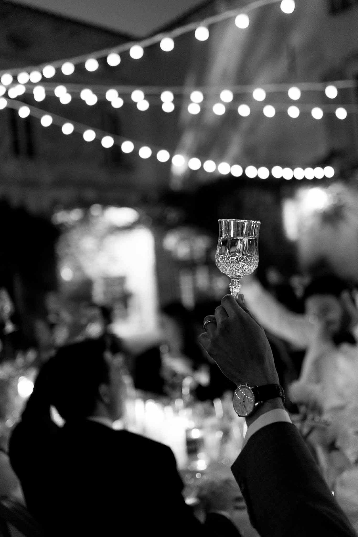 Black and white close-up of a crystal champagne glass raised in a toast during a wedding reception with festoon lights