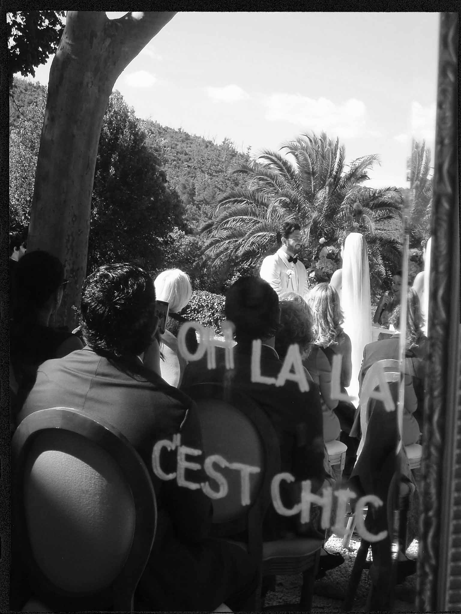Black-and-white view of outdoor wedding ceremony through glass with hand-lettered French text, guests seated facing altar