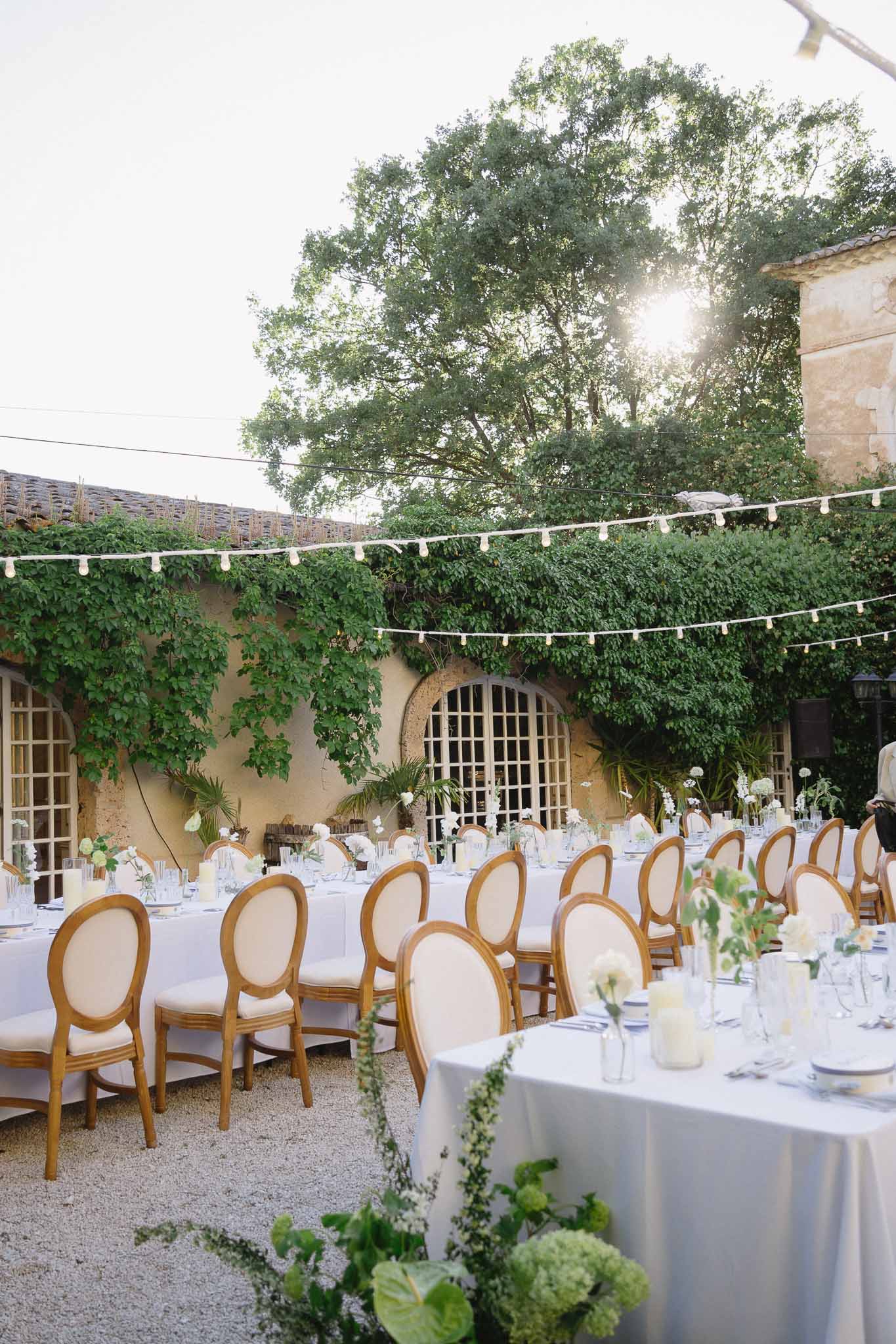 Long tables in grey-blue linen with white and green bud vases under globe lights in ivy-walled courtyard