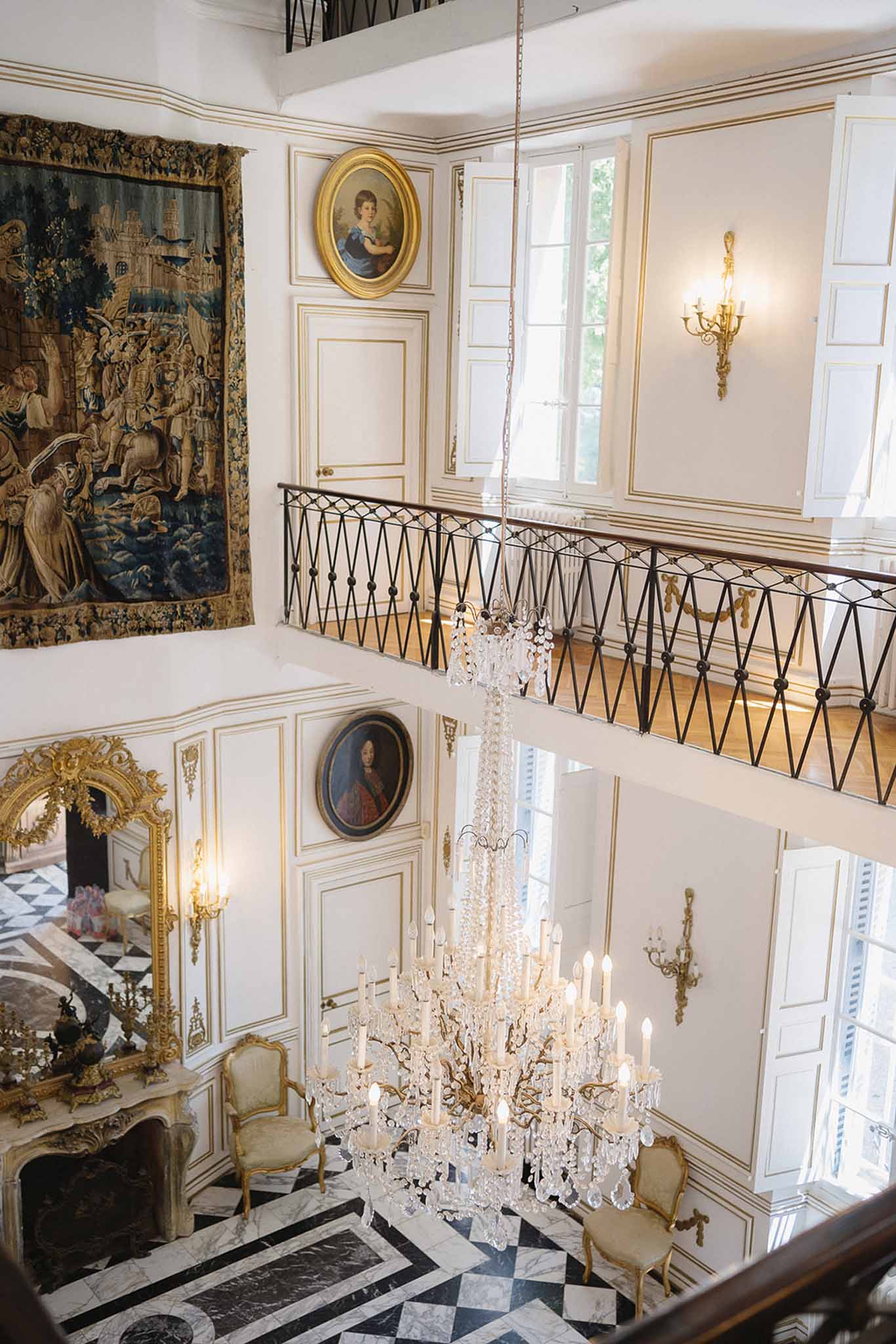 Double-height chateau entrance hall with crystal chandelier, marble floor, tapestry, and gilt furniture
