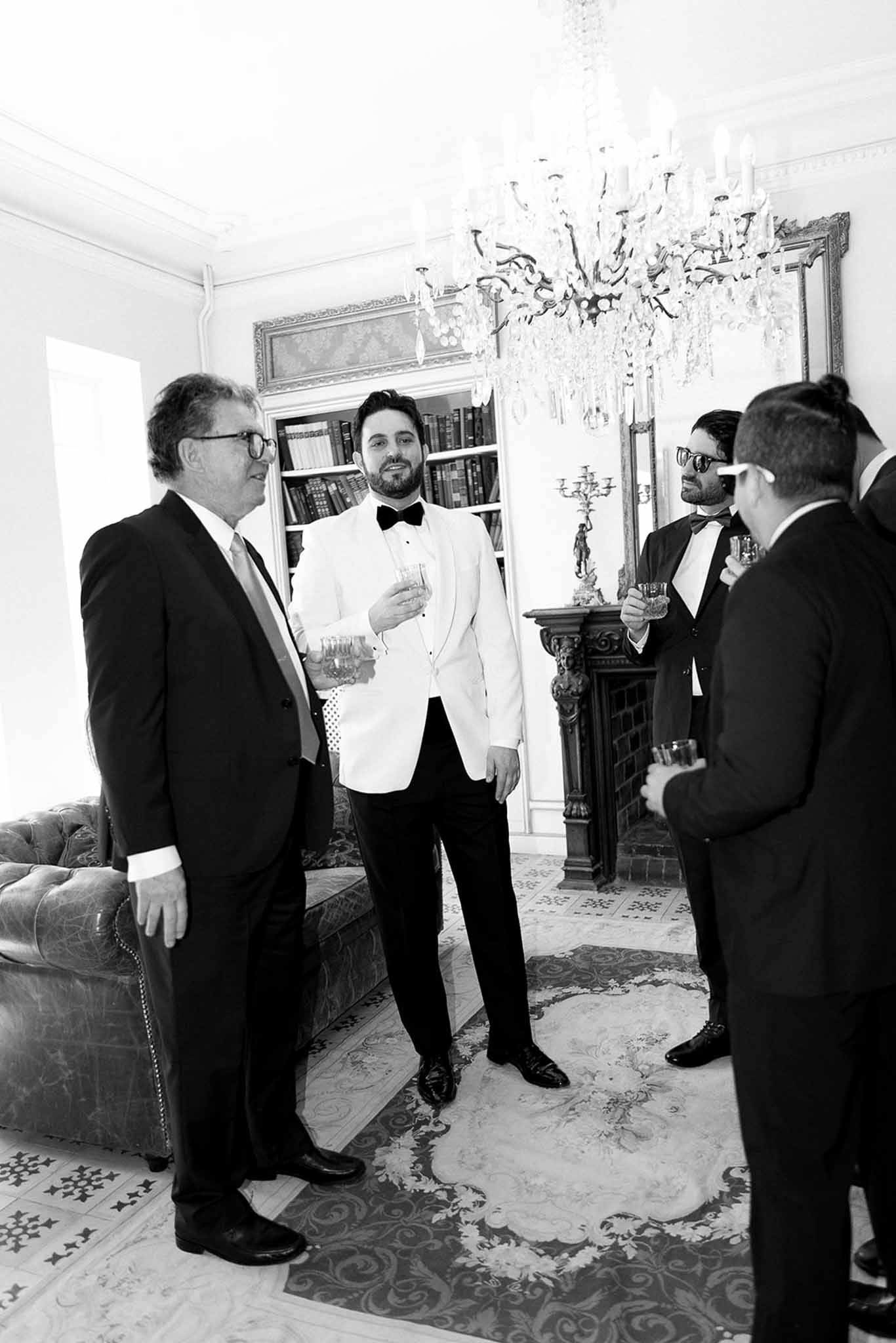 Black and white groom in white dinner jacket with groomsmen in chateau salon with chandelier fireplace and Chesterfield sofa
