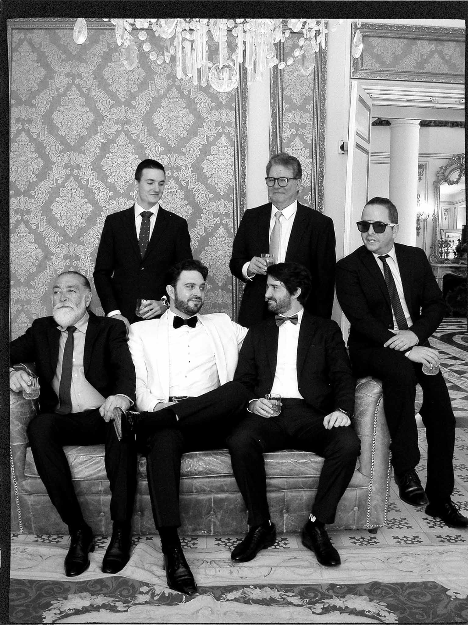 Black and white group of six men with groom in white dinner jacket on damask sofa in ornate salon