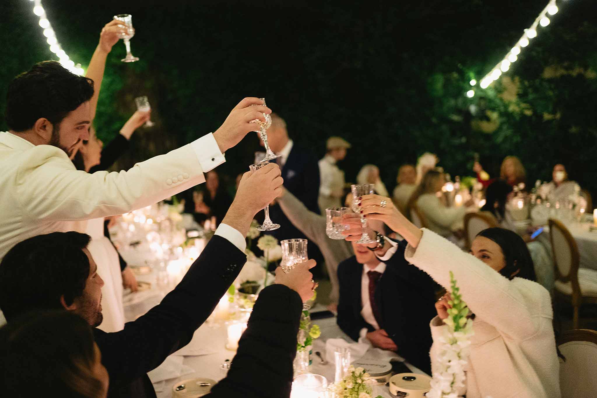 Guests raising champagne glasses during toast at candlelit outdoor evening reception