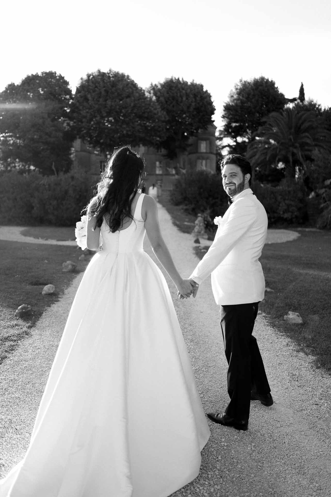 Bride and groom walking away hand-in-hand on gravel path toward manor house in black and white