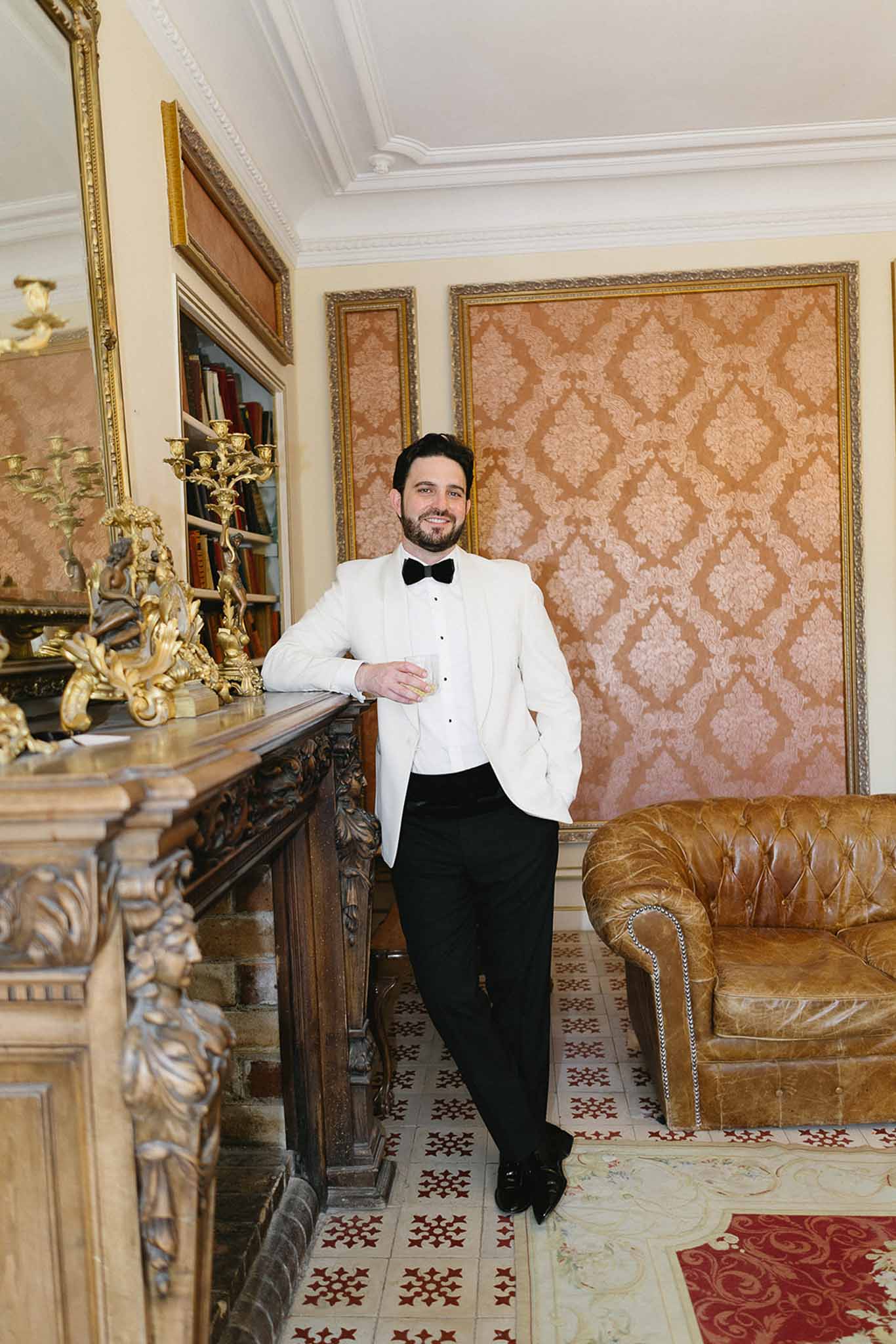 Groom in white dinner jacket and black trousers leaning against ornate fireplace in chateau interior room