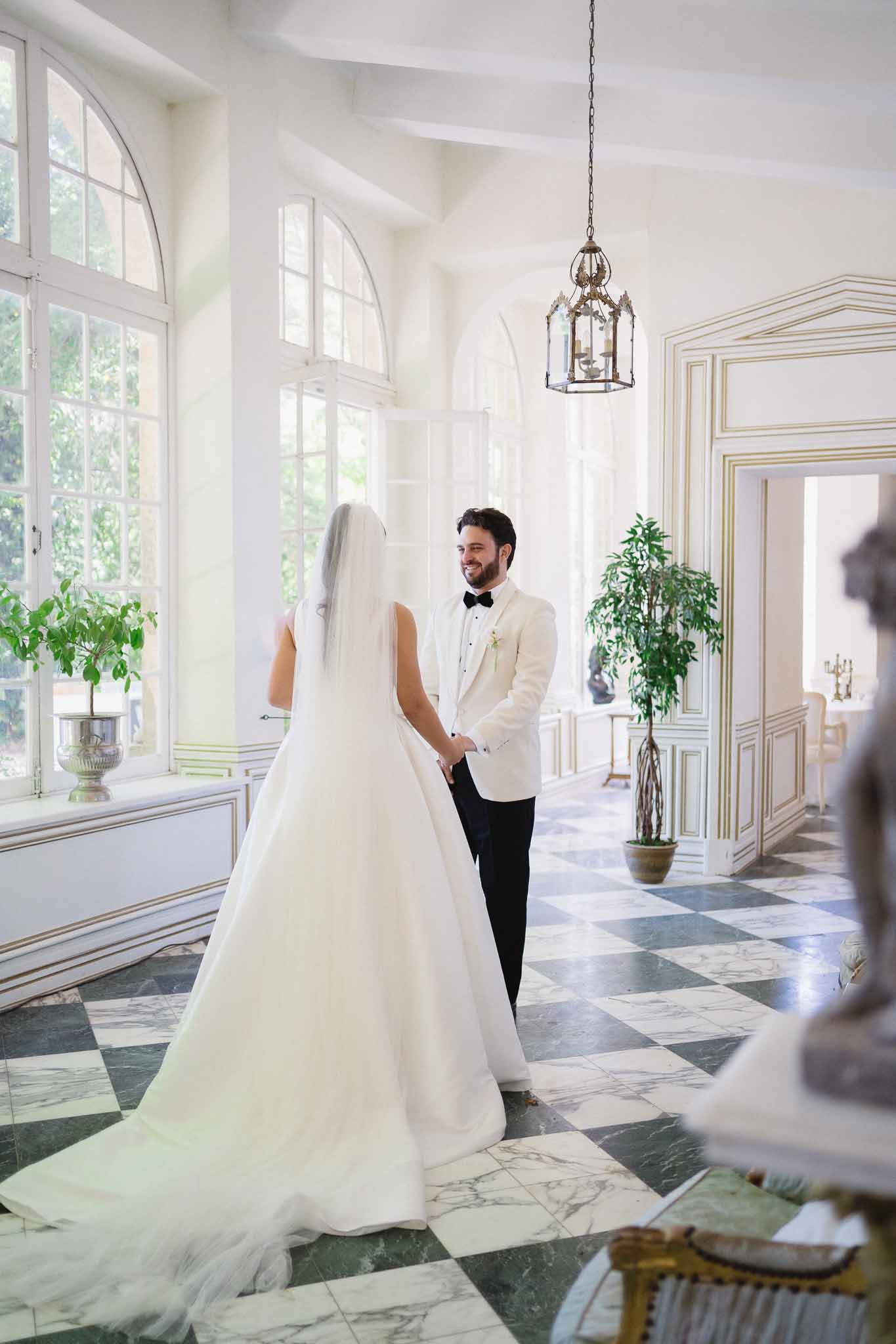 First look on black and white marble floor with groom smiling at veiled bride in gold-trimmed boiserie hall