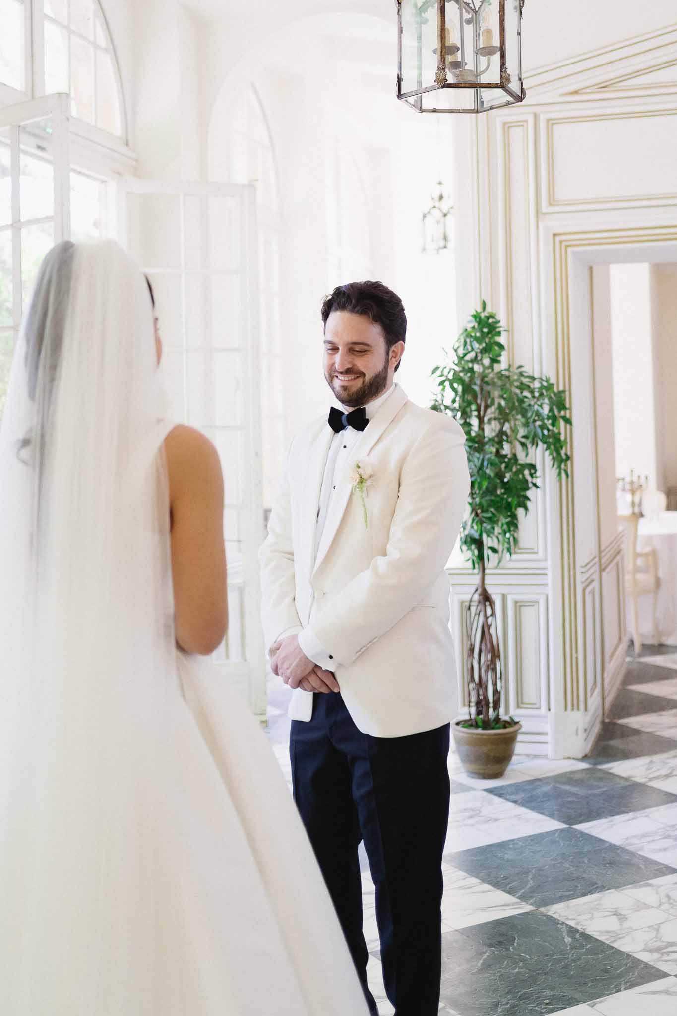 Groom in ivory dinner jacket smiling at bride with cathedral veil in marble chequerboard hallway with glass lantern
