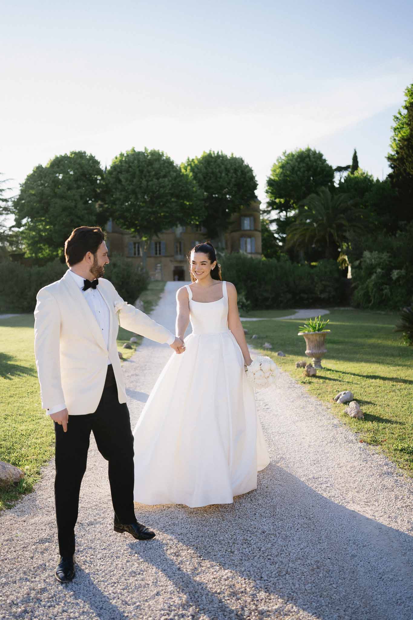Groom in white dinner jacket leading bride with white bouquet along chateau driveway in golden hour