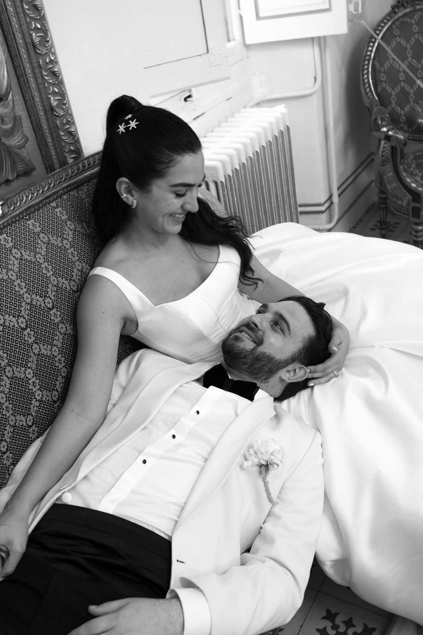 Black-and-white portrait of groom lying in bride's lap on ornate sofa in classic French venue interior