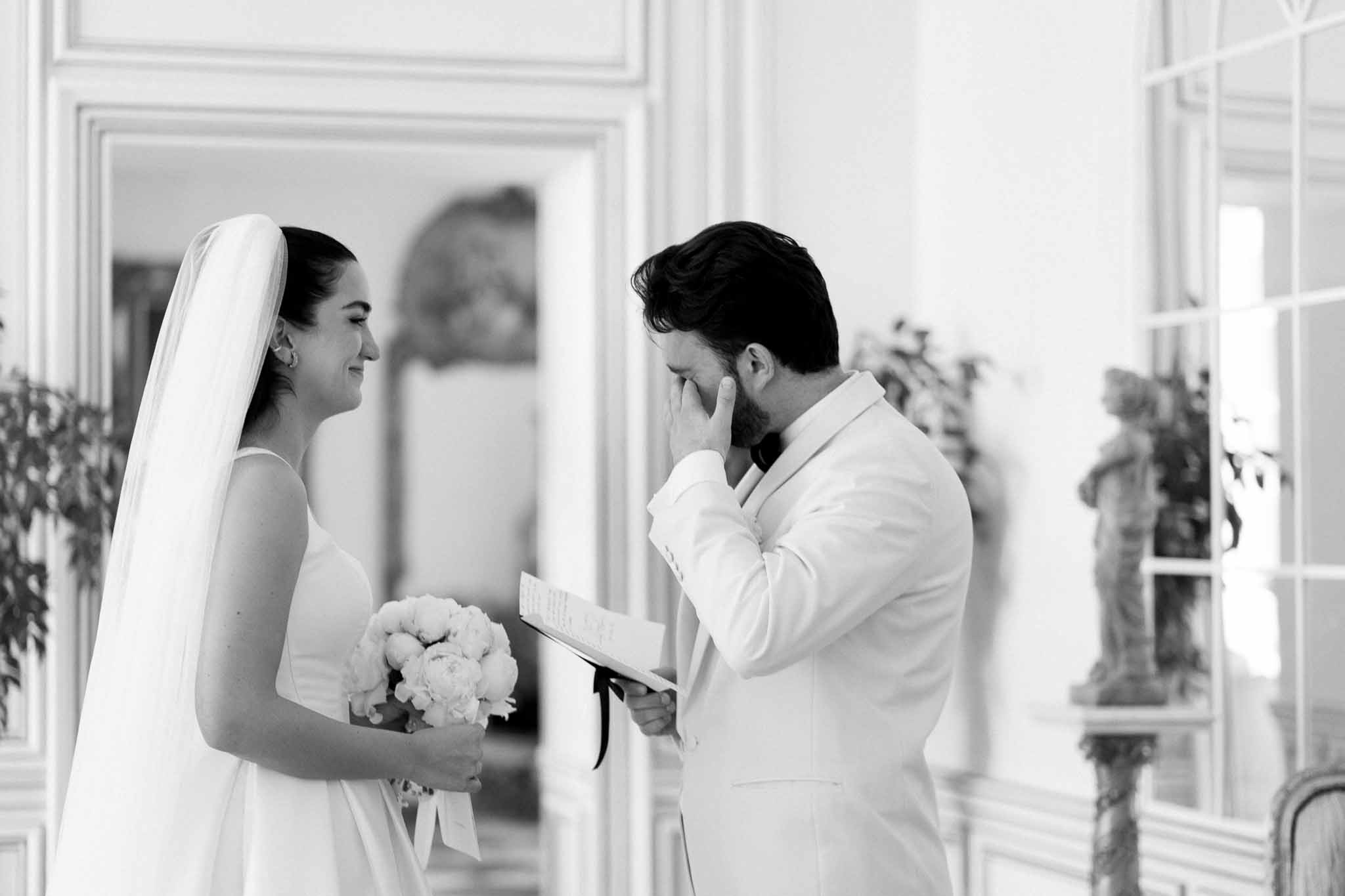 Black-and-white shot of groom reading vows while covering face emotionally as bride smiles holding peony bouquet indoors