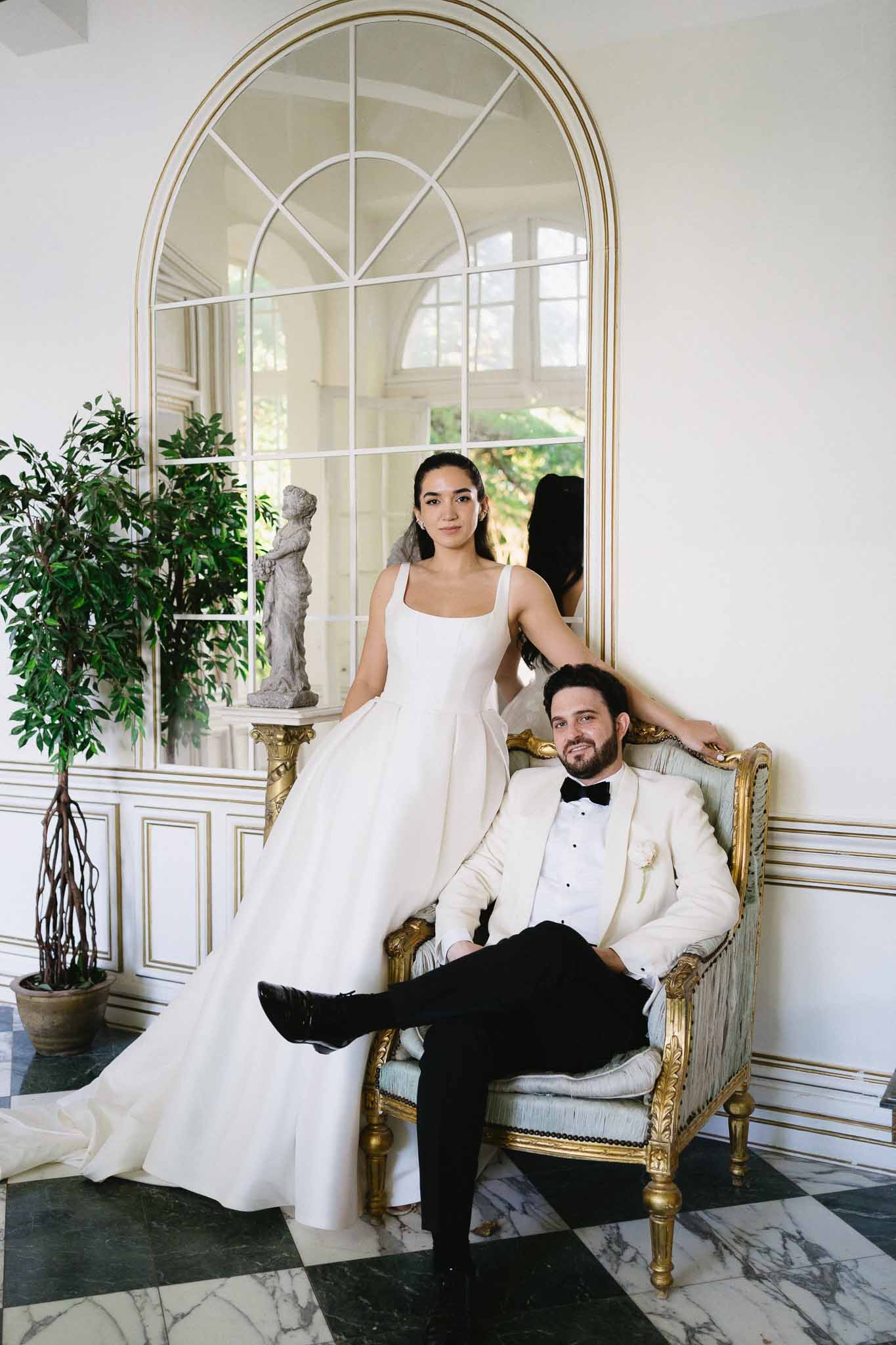 Groom seated on grey silk settee with bride standing behind in ballgown before gilt mirror on marble floor