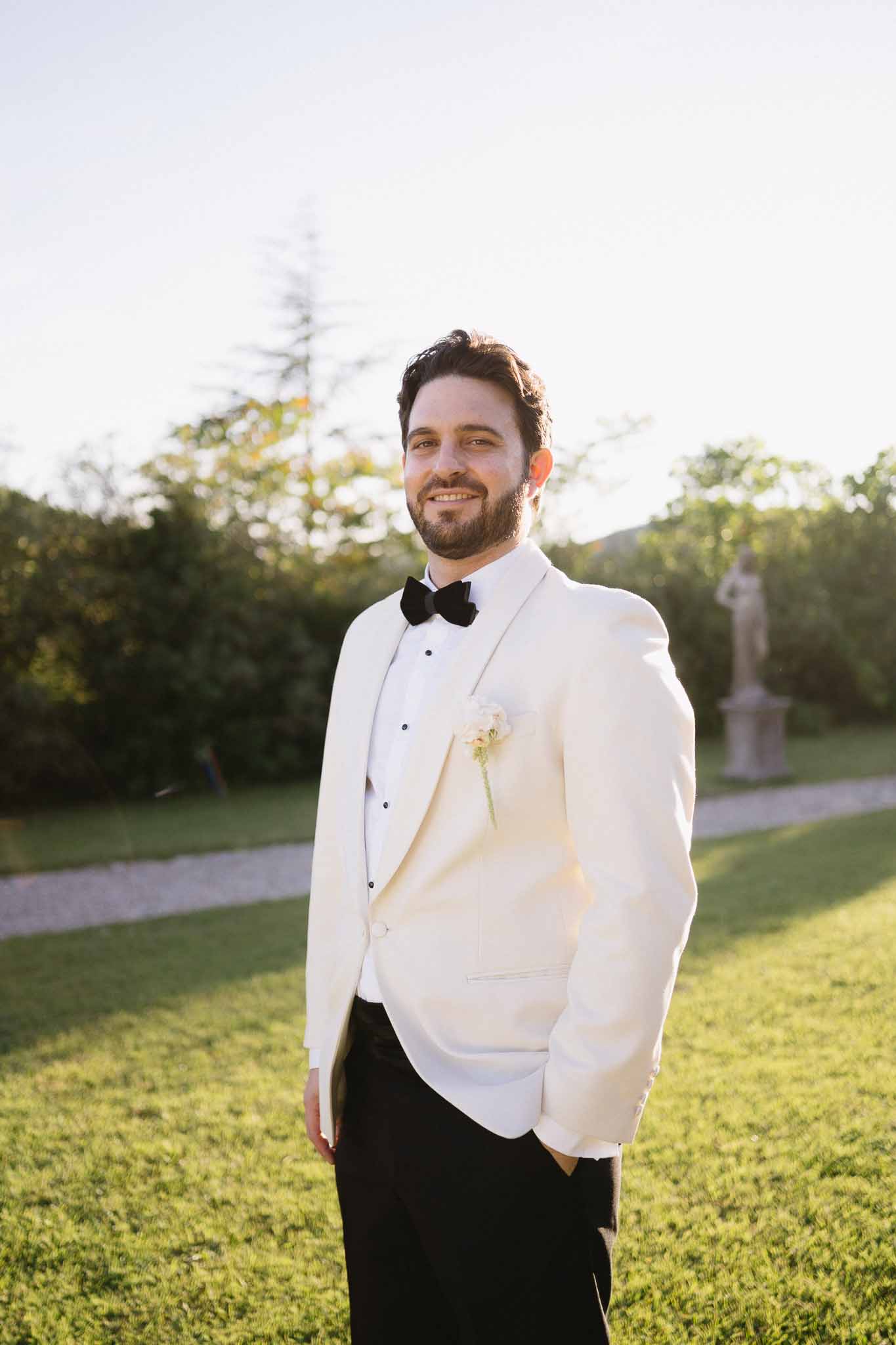 Groom in cream dinner jacket with black bow tie and white boutonniere at golden hour in chateau garden