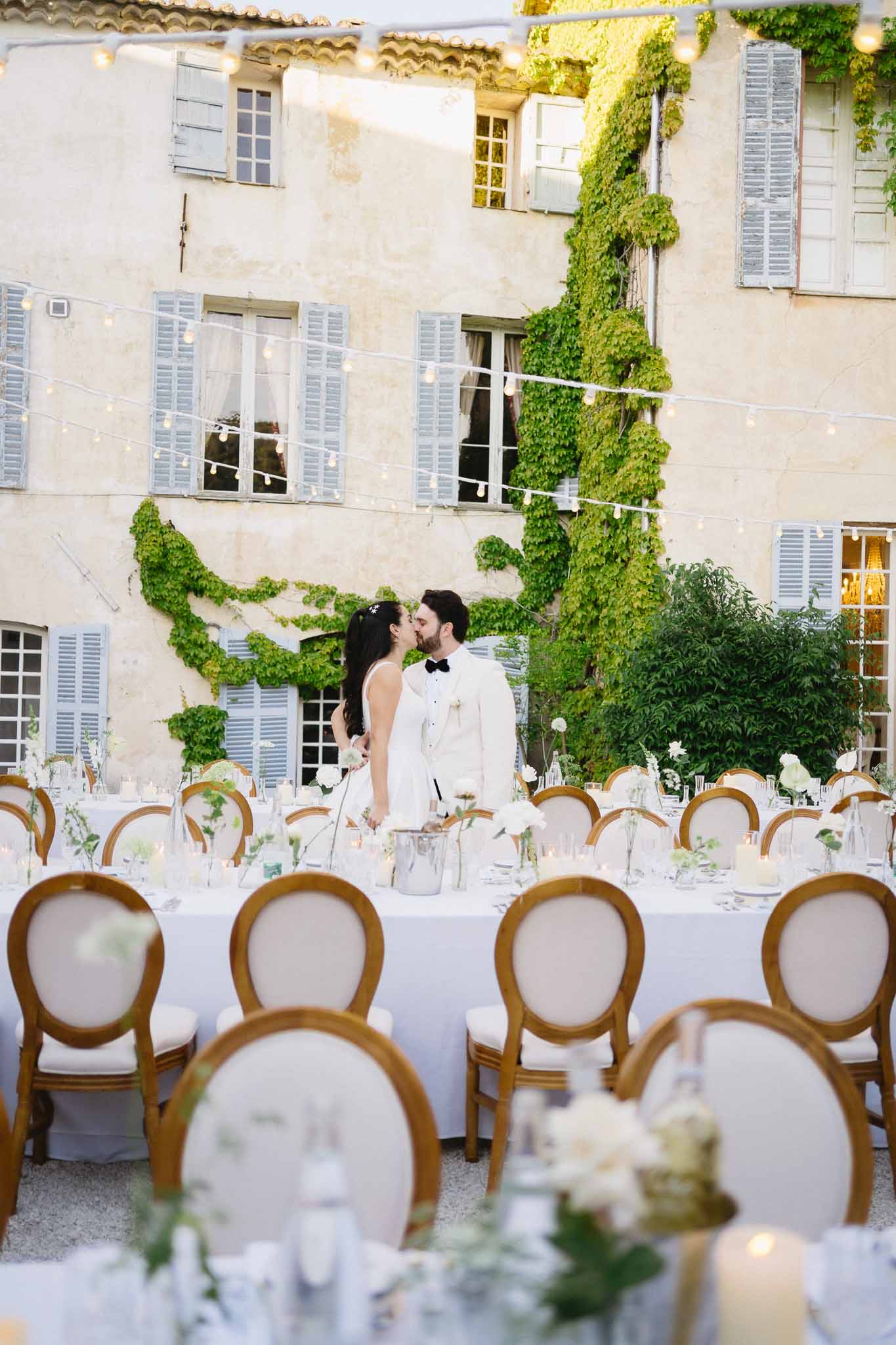 Couple kissing at candlelit courtyard reception with white bud vases and gold medallion chairs before Provencal mas