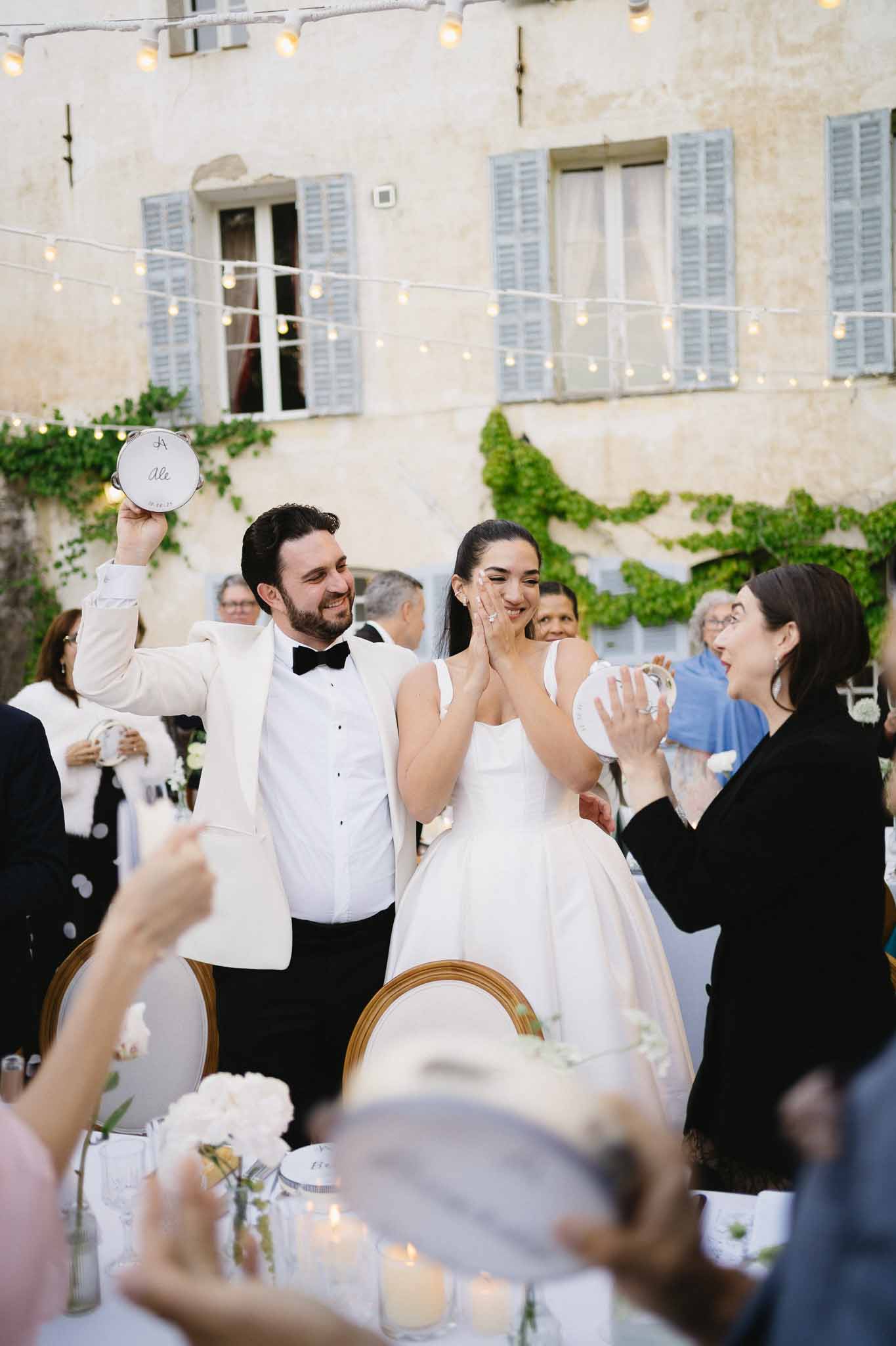Bride and groom shaking tambourines at sweetheart table in fairy-lit French courtyard reception