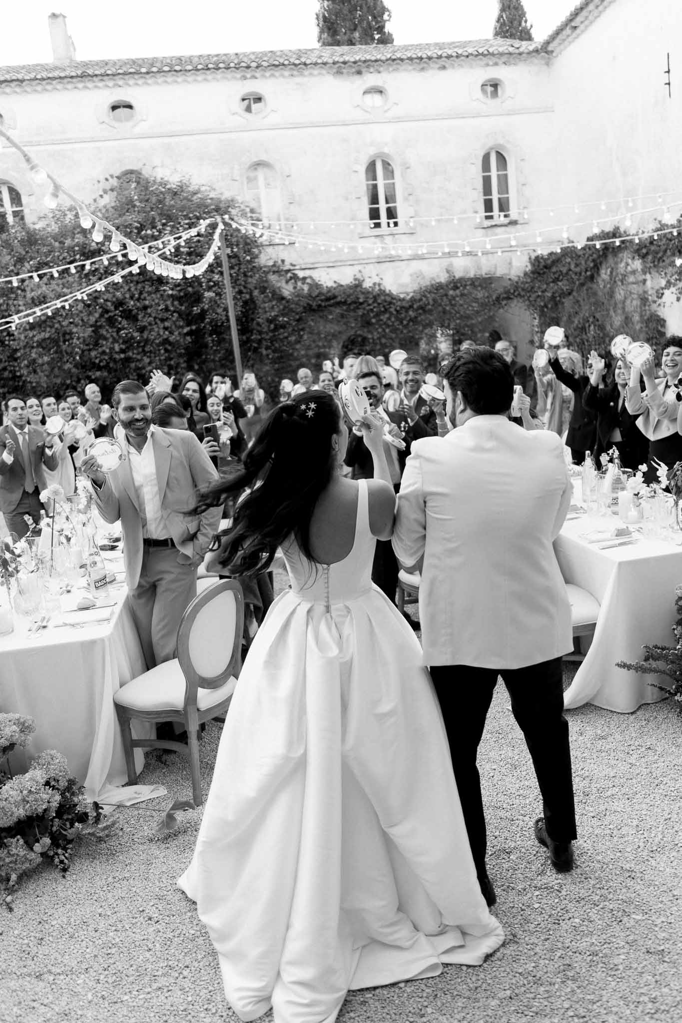 Bride and groom entering outdoor courtyard reception with tambourines as guests cheer under festoon lights