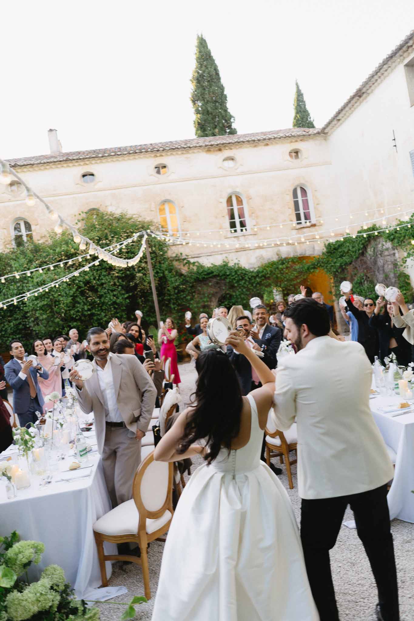 Couple with tambourines as guests celebrate at candlelit courtyard reception with gold chairs and string lights