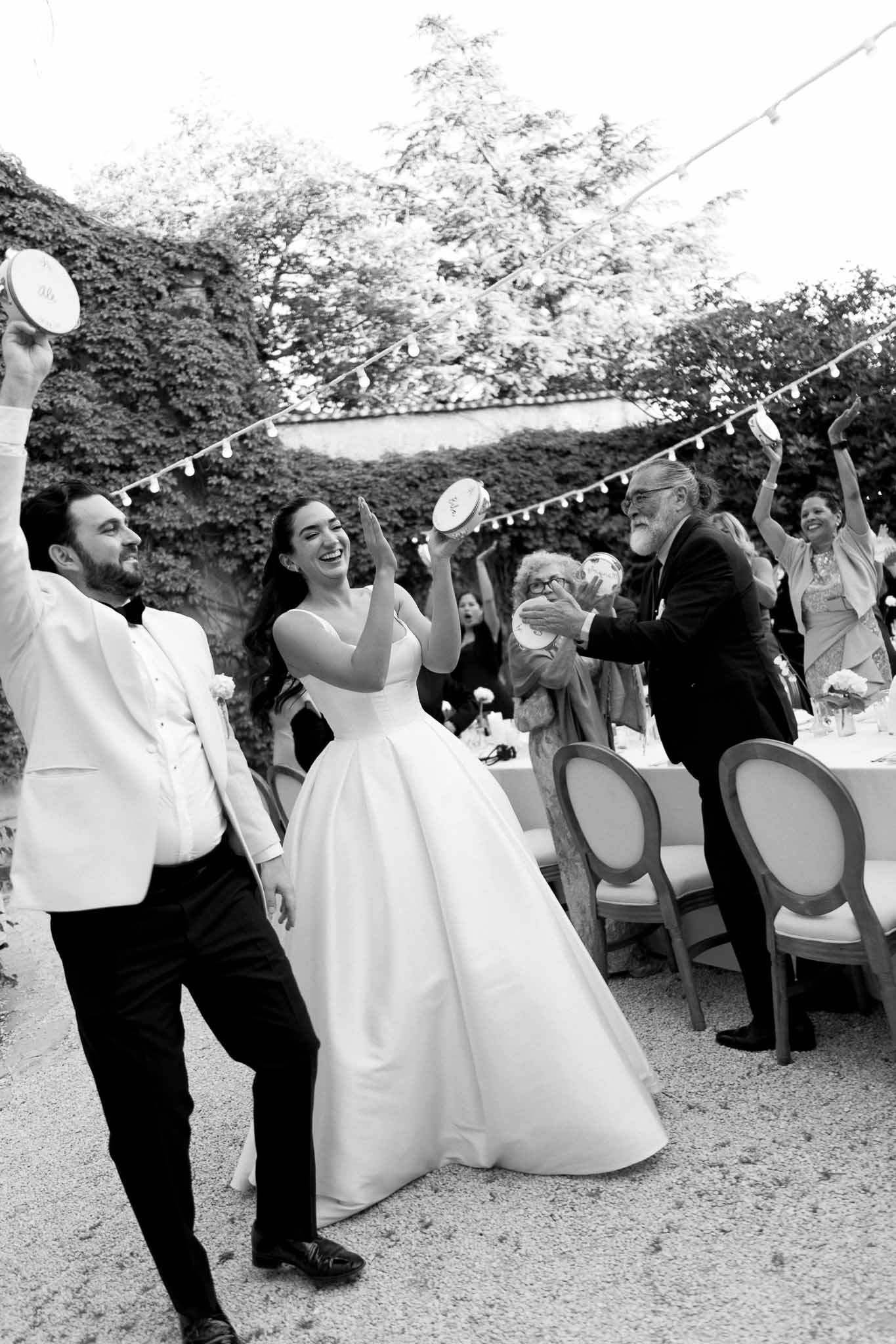 Black and white couple playing tambourines with dancing guests under string lights in courtyard
