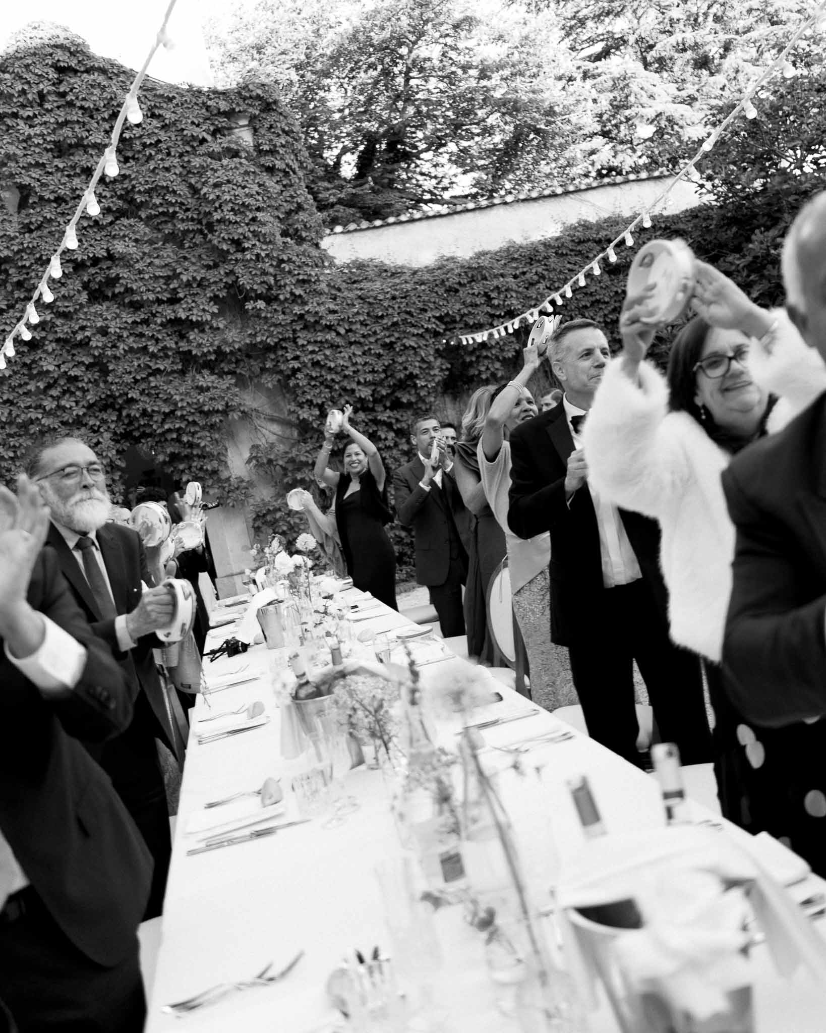 Guests wave napkins in celebratory toast at long table with globe lights and bunting in B&W