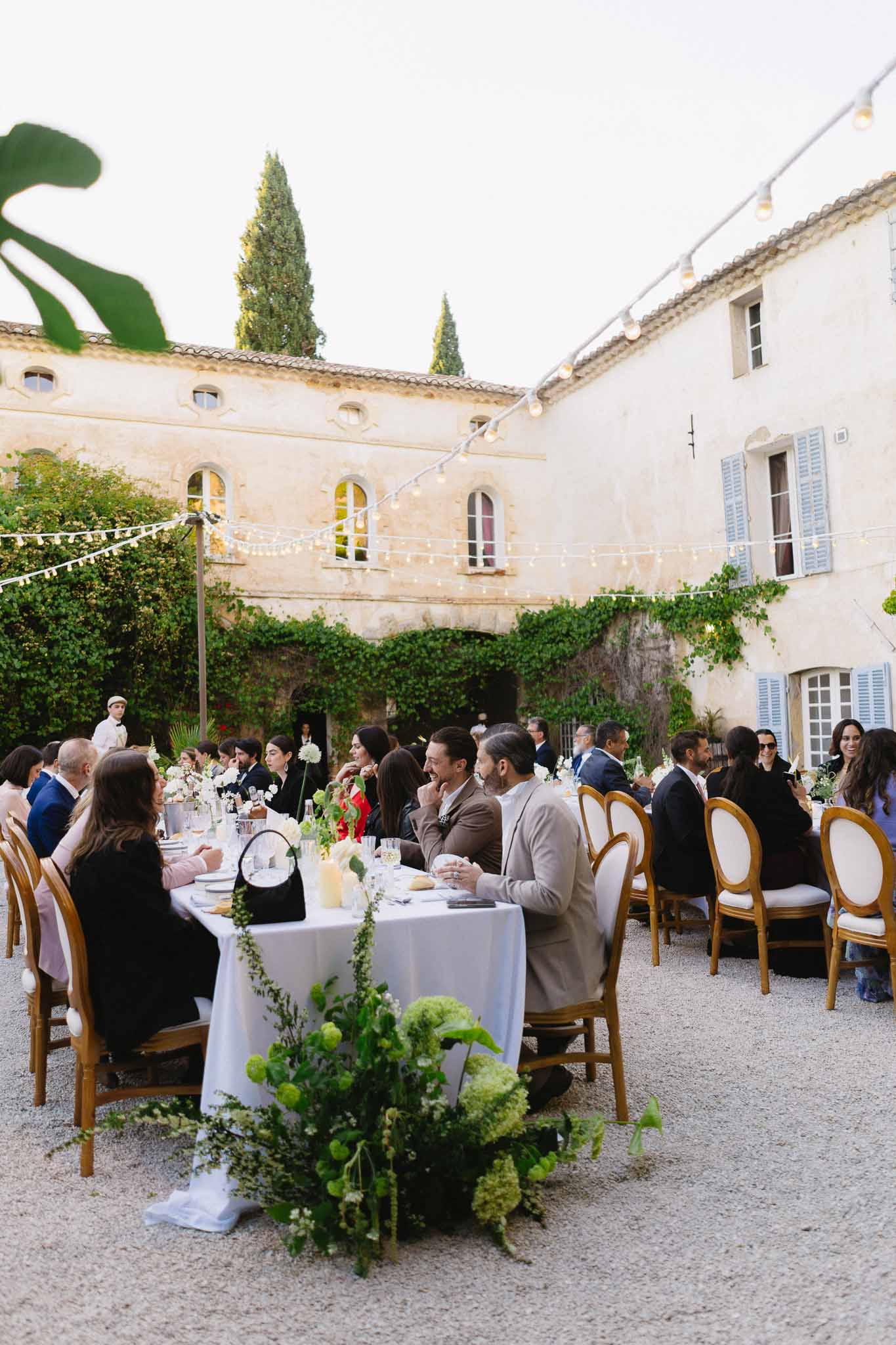 Forty guests at long tables with trailing viburnum centerpieces under Edison bulb lights in mas courtyard