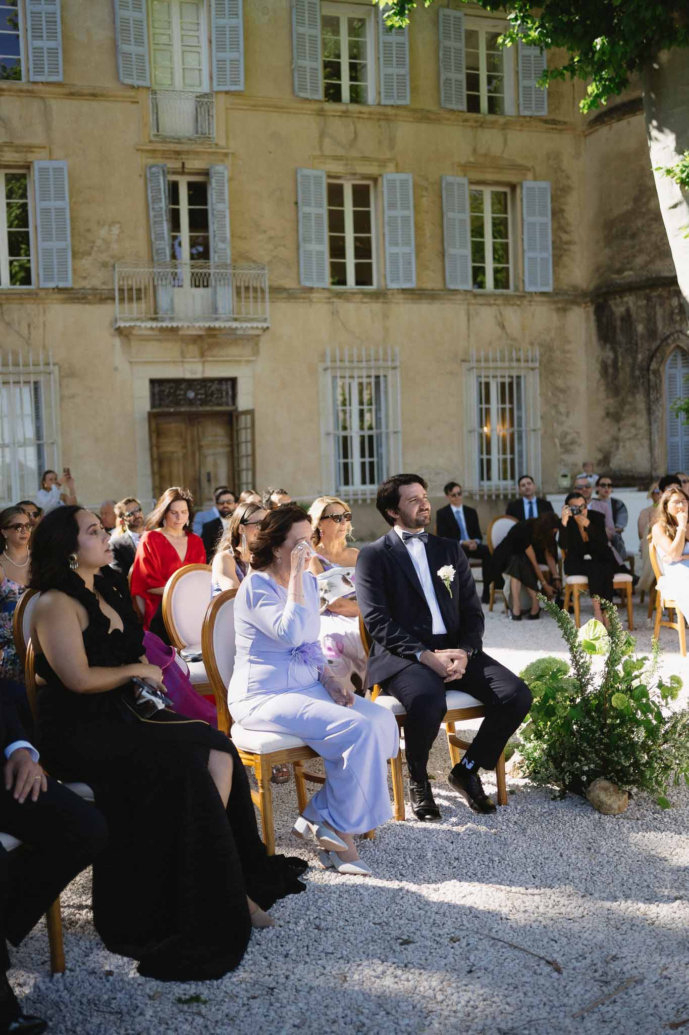 Thirty guests on gold Chiavari chairs watching ceremony at yellow chateau with wrought-iron balconies