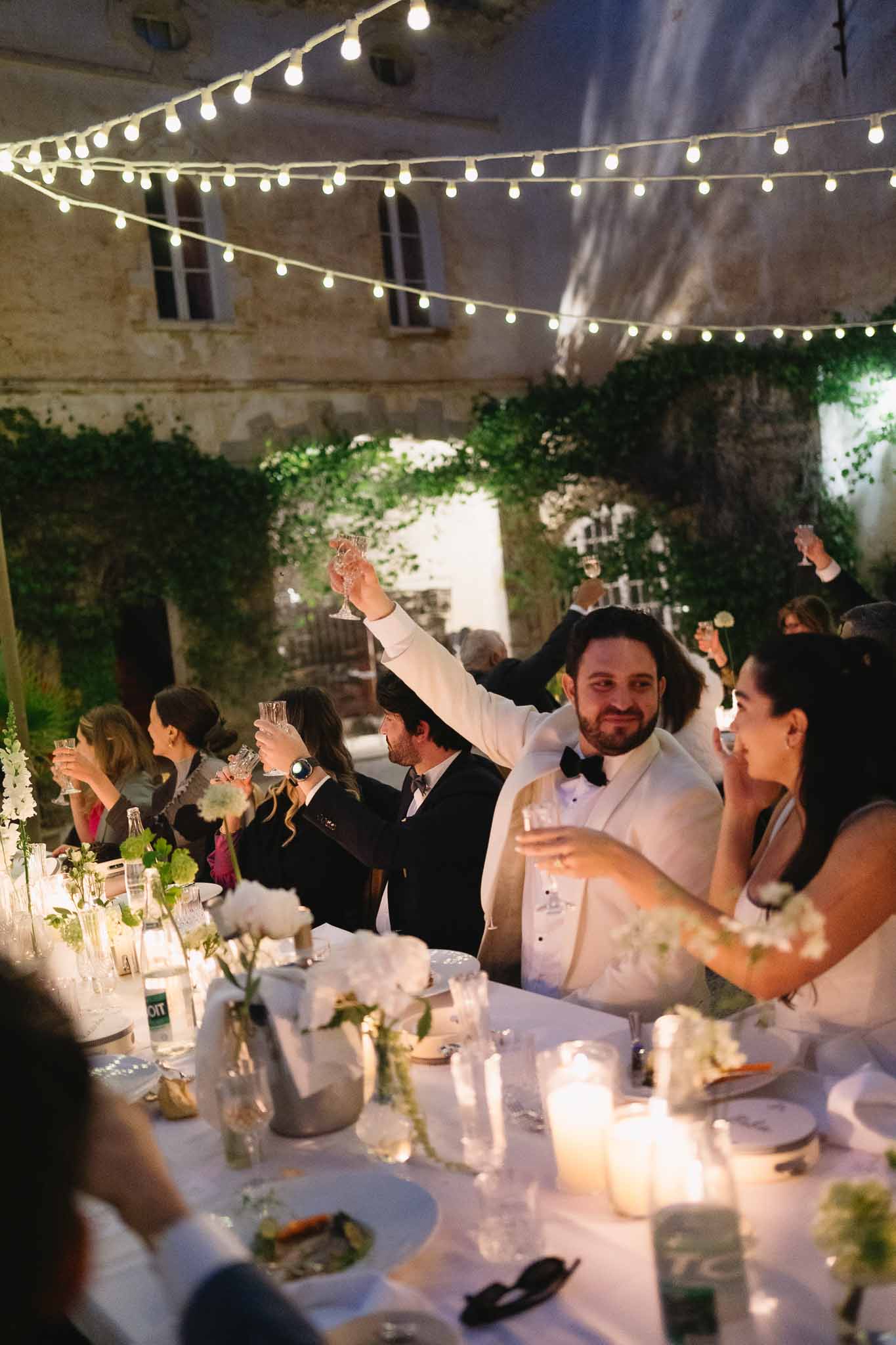 Groom in white dinner jacket raising champagne coupe at string-lit ivy courtyard dinner with white peonies