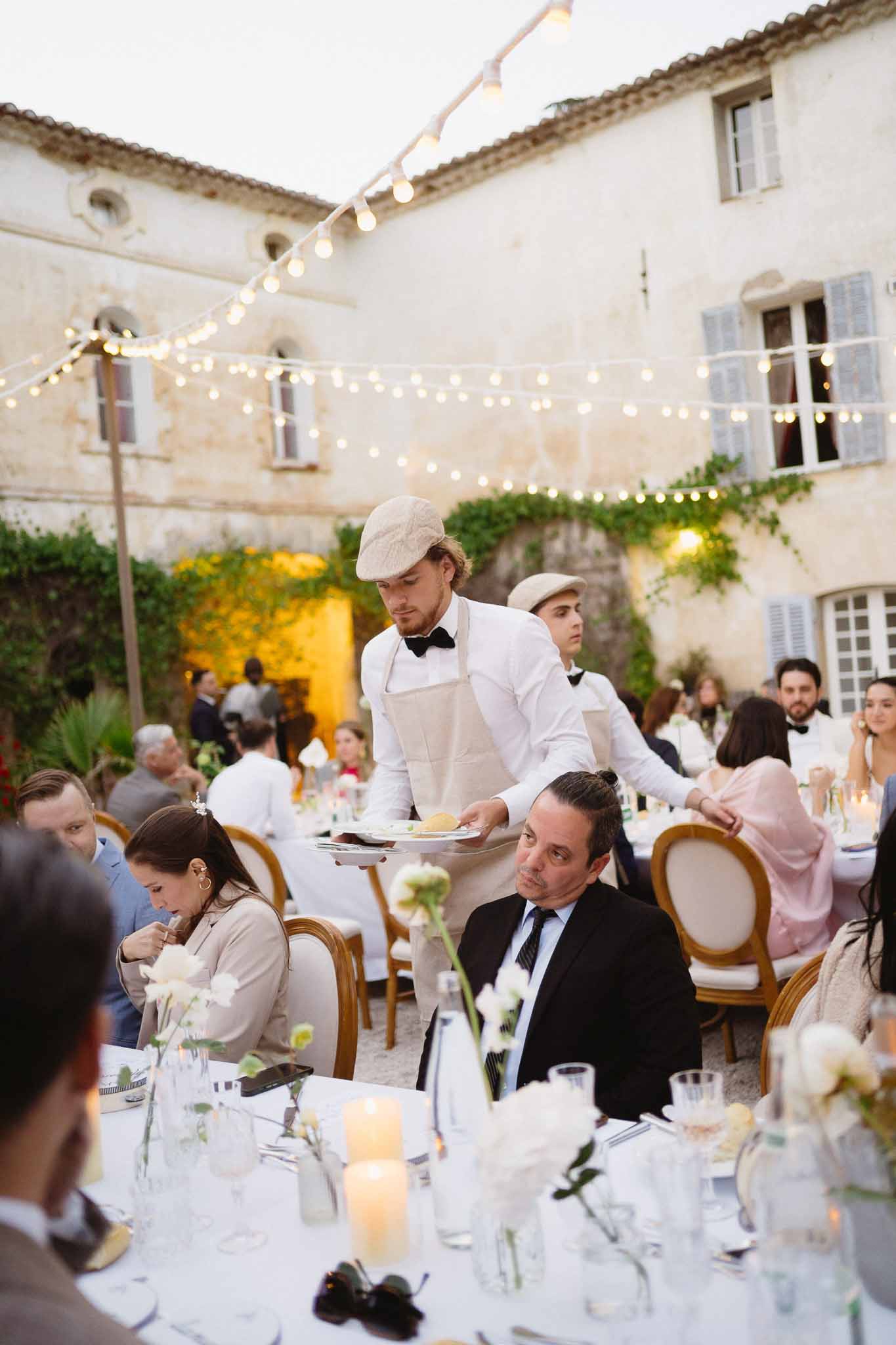 Waiters serving dusk reception with white roses, pillar candles, and gold Louis chairs under fairy lights