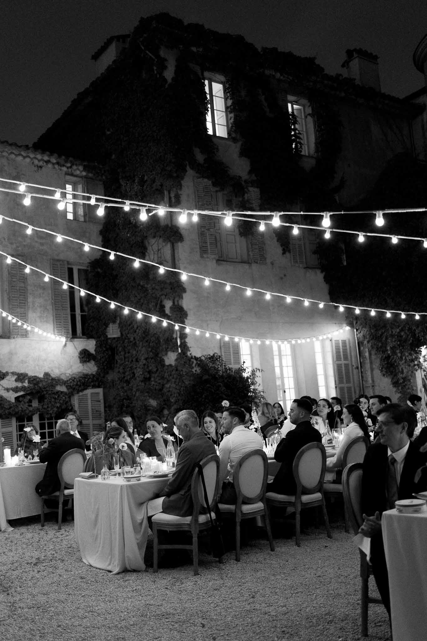 Black-and-white evening reception in ivy-covered chateau courtyard with bistro lights and seated guests