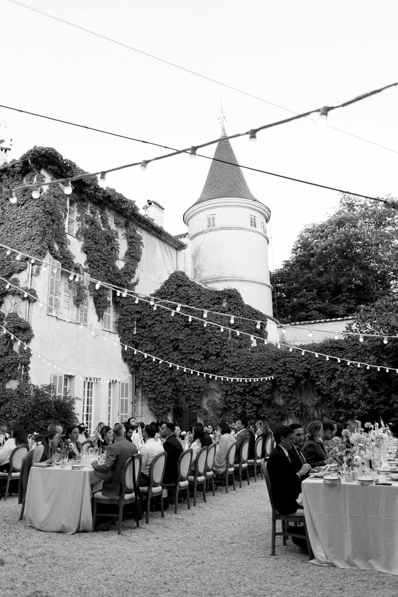 Black and white wide shot of outdoor reception dinner with guests at long tables in chateau courtyard with fairy lights