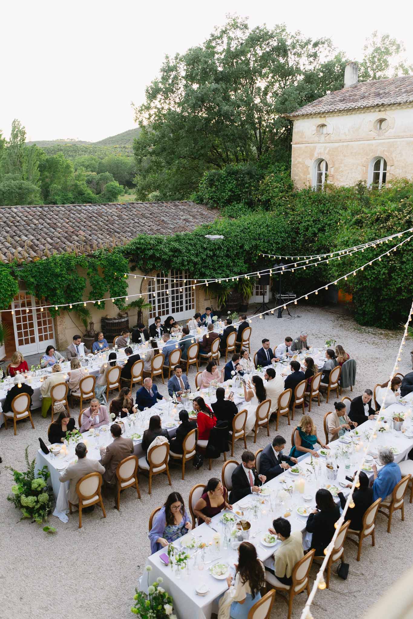 Sixty-five guests at U-shaped tables under globe lights in ivy-covered stone mas courtyard at night