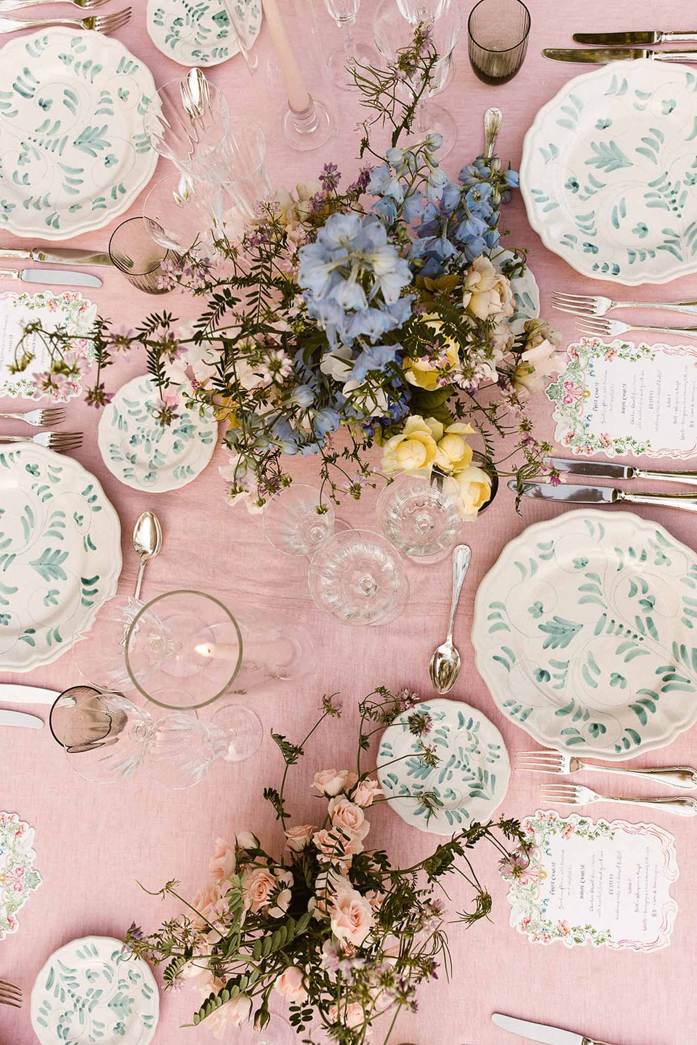 Overhead dusty pink table with botanical painted plates, blue delphiniums, yellow roses, and die-cut menus