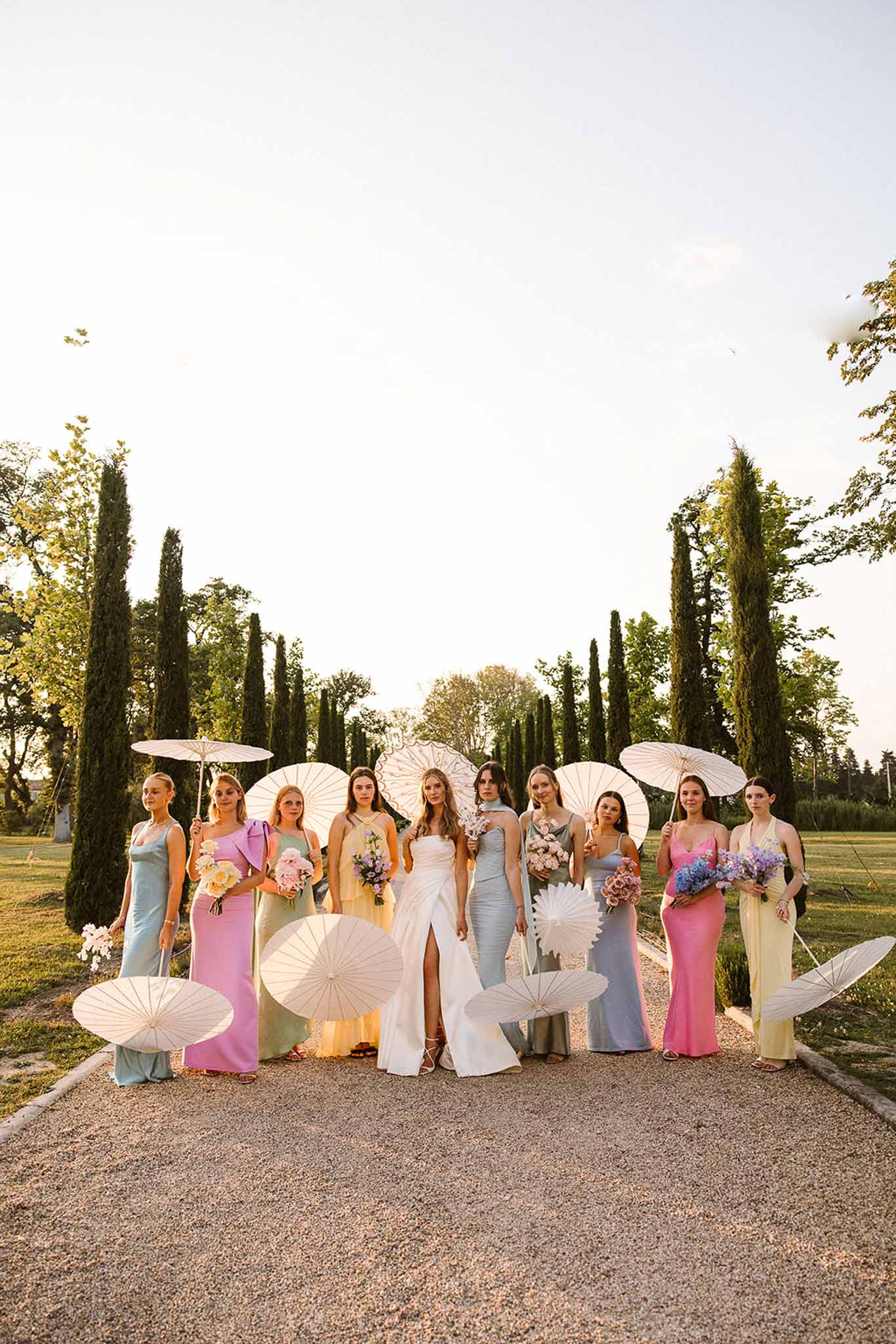 Bride with nine bridesmaids in mismatched pastel dresses holding white parasols on a cypress-lined driveway at golden hour