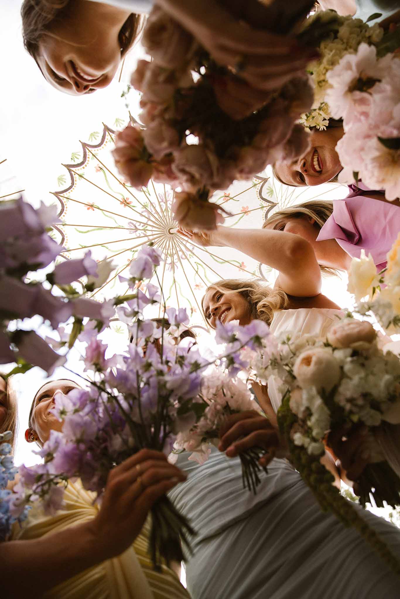Low-angle shot of bride and four bridesmaids in pastel gowns holding garden-style bouquets with floral parasol