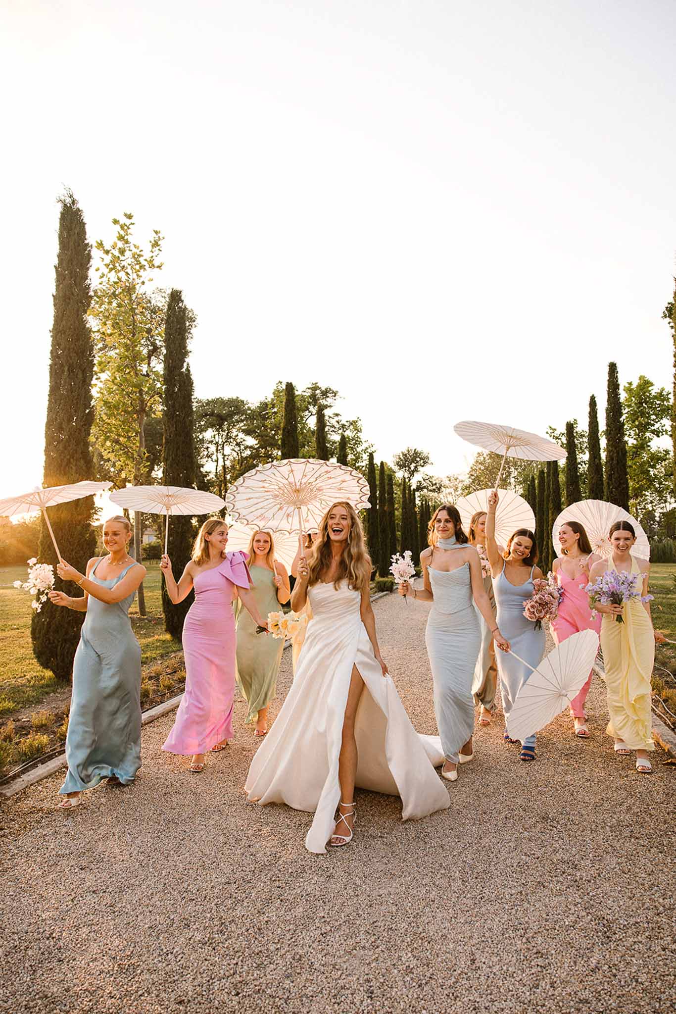 Bride and eight bridesmaids in mismatched pastel dresses with paper parasols on cypress path at golden hour