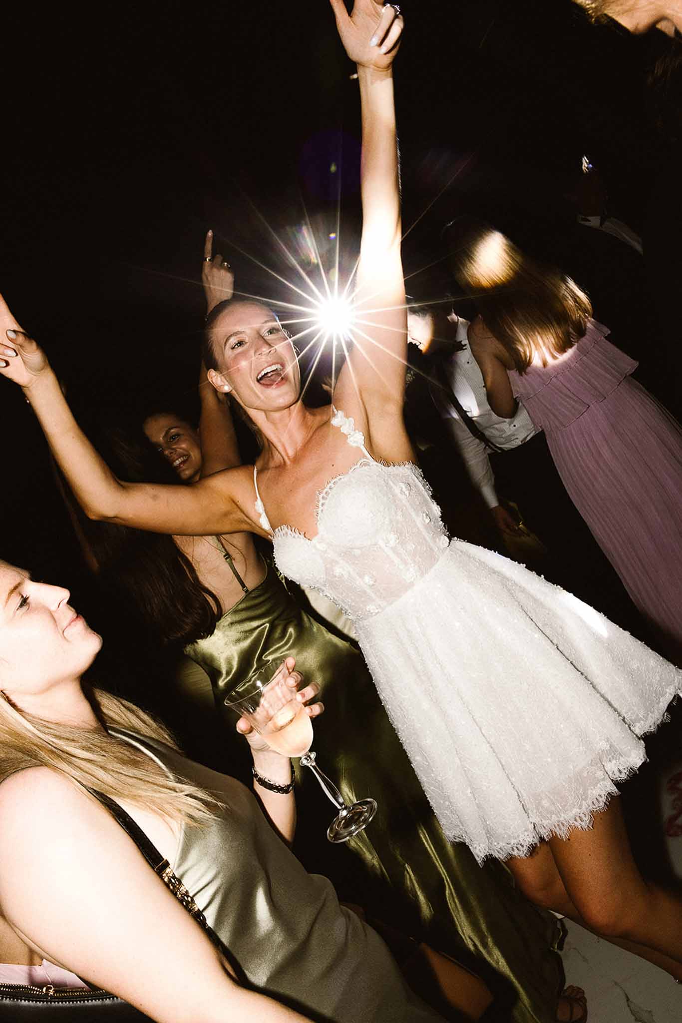Bride dancing in white lace mini dress with champagne flute surrounded by guests under starburst light