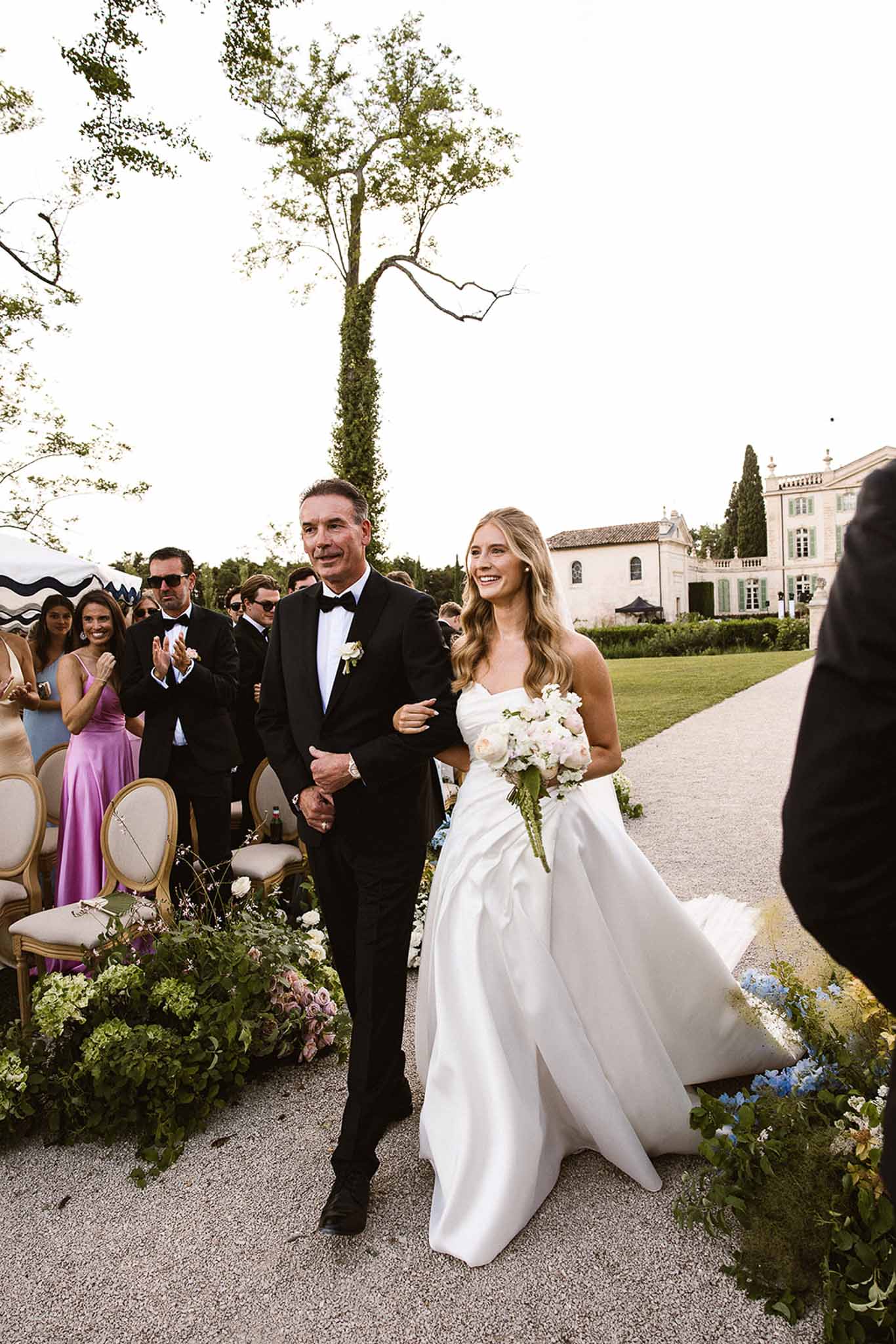 Bride walking down gravel aisle with father past blue delphinium and blush rose arrangements