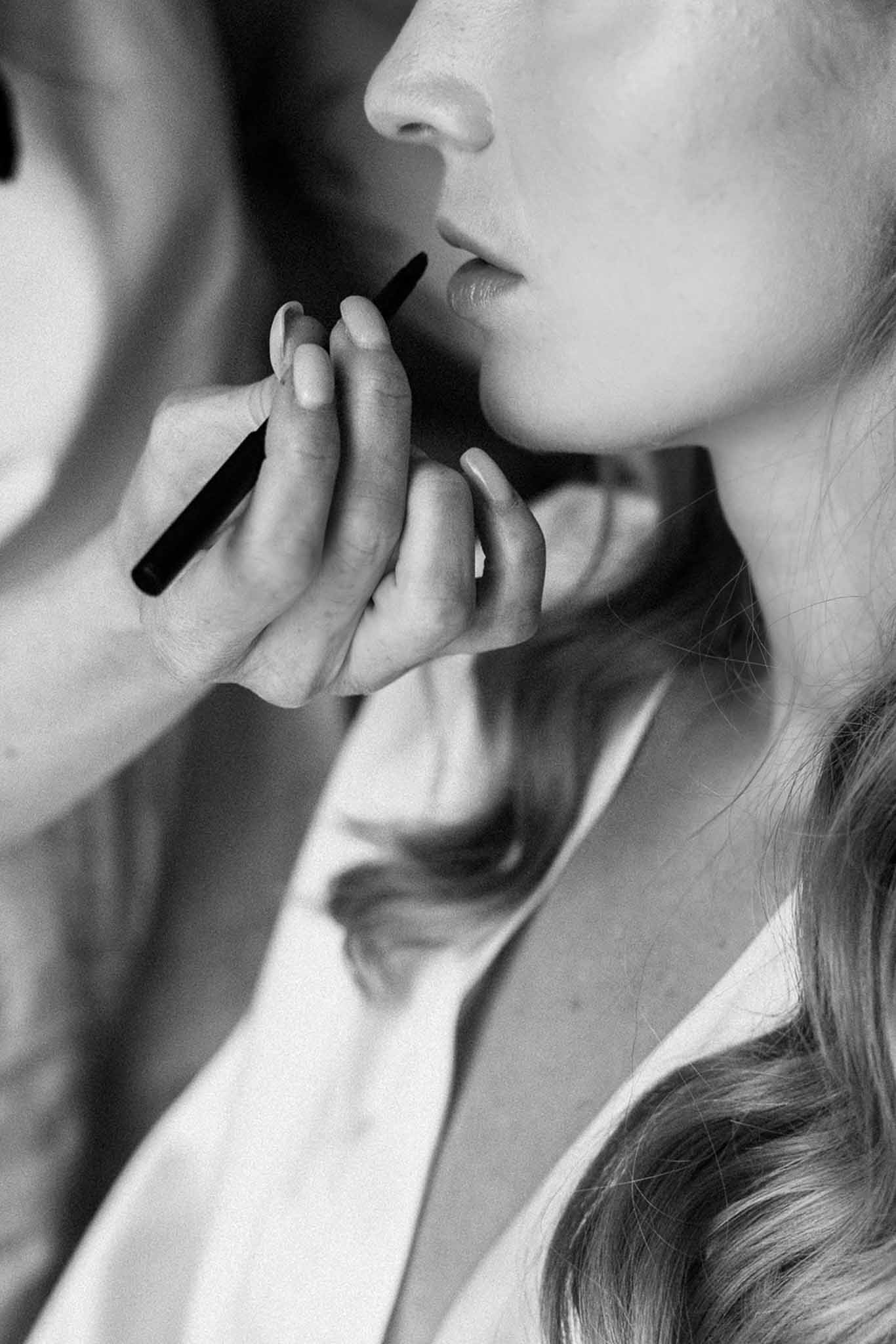 Black and white close-up of makeup artist applying lip liner to bride in white robe