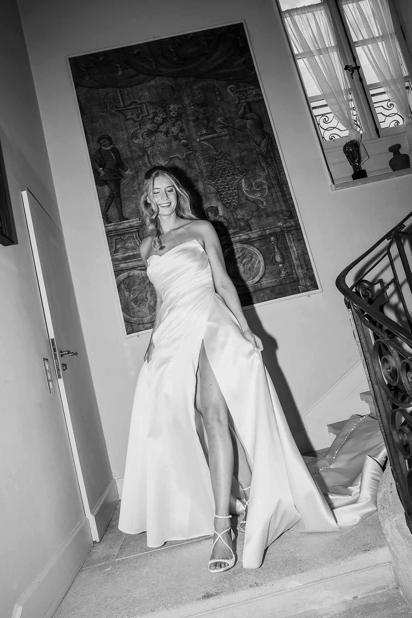 Black and white bride laughing and lifting ruched satin gown on stone staircase with wrought-iron railing and mural
