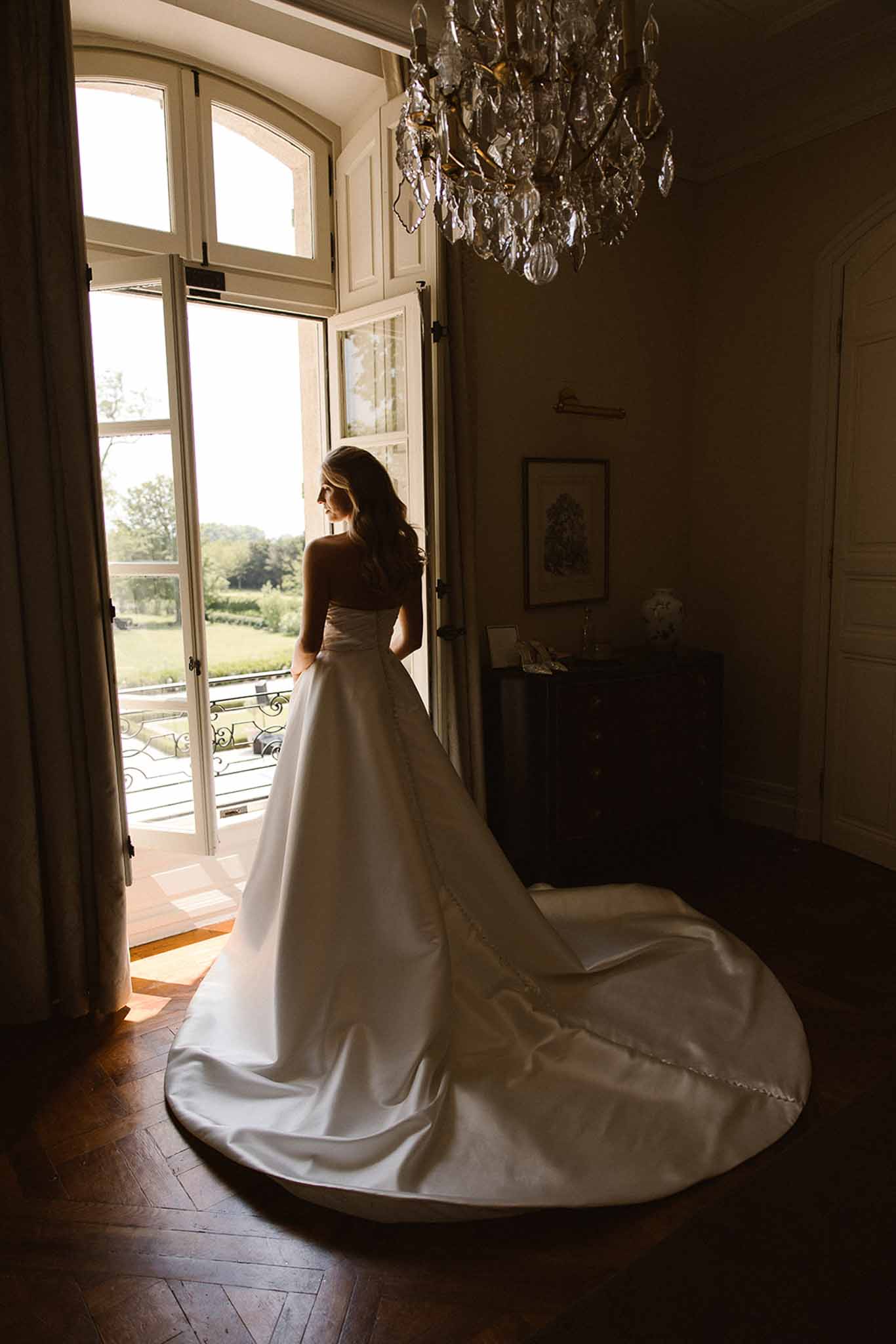 Bride in strapless satin ball gown with cathedral train silhouetted at open French doors of chateau