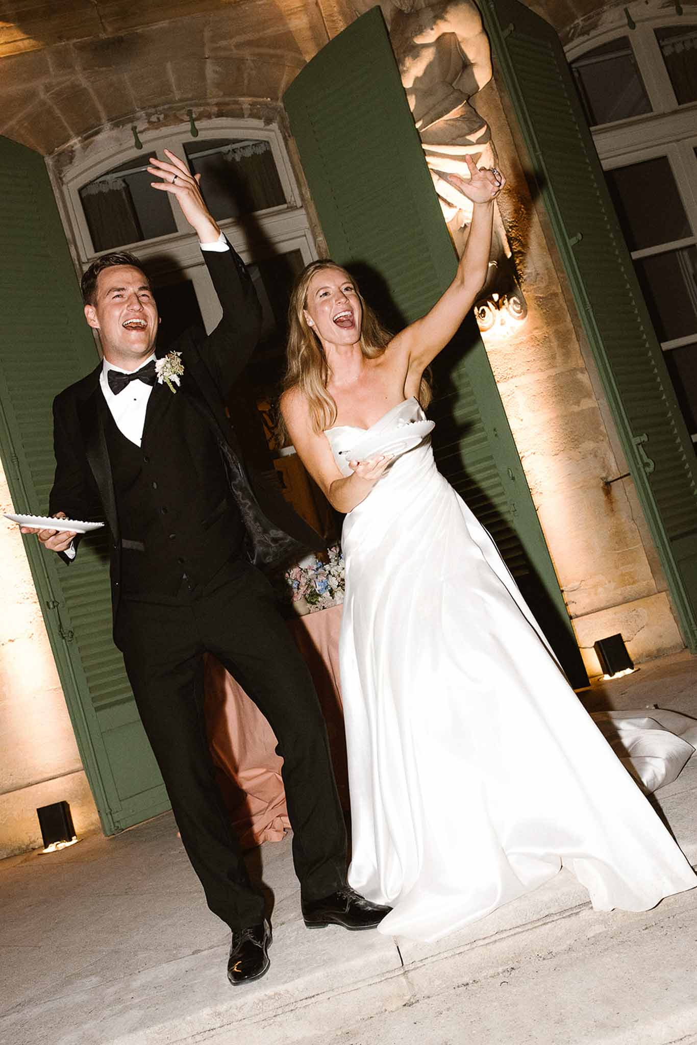 Bride in strapless ballgown and groom in tuxedo raising arms at chateau doorway with sage shutters at night