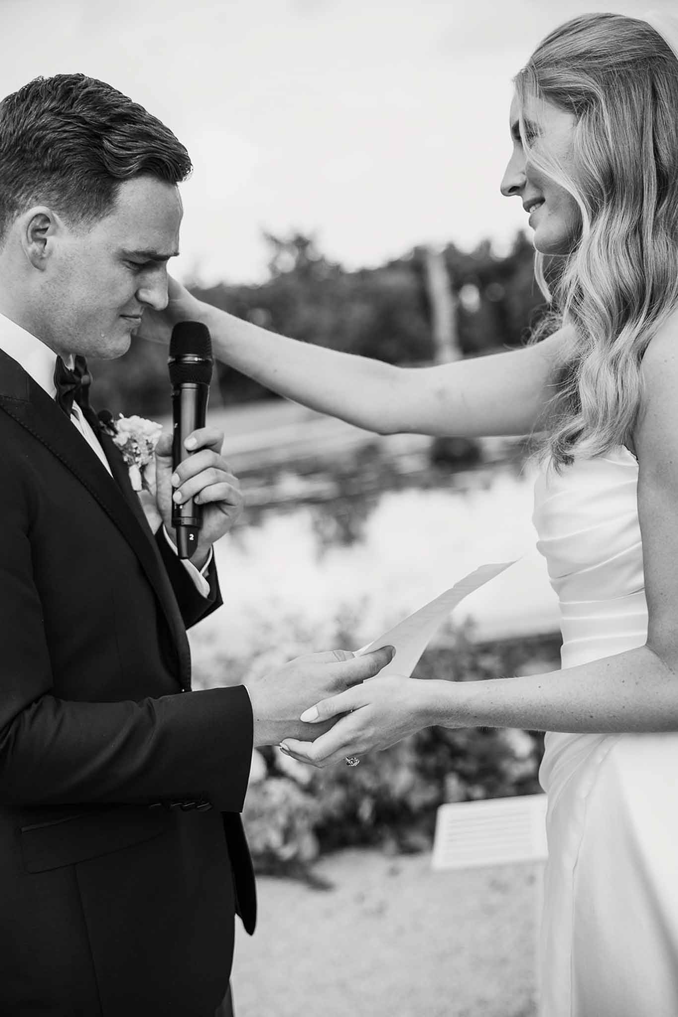 Black and white close-up of emotional groom reading vows as bride touches his face