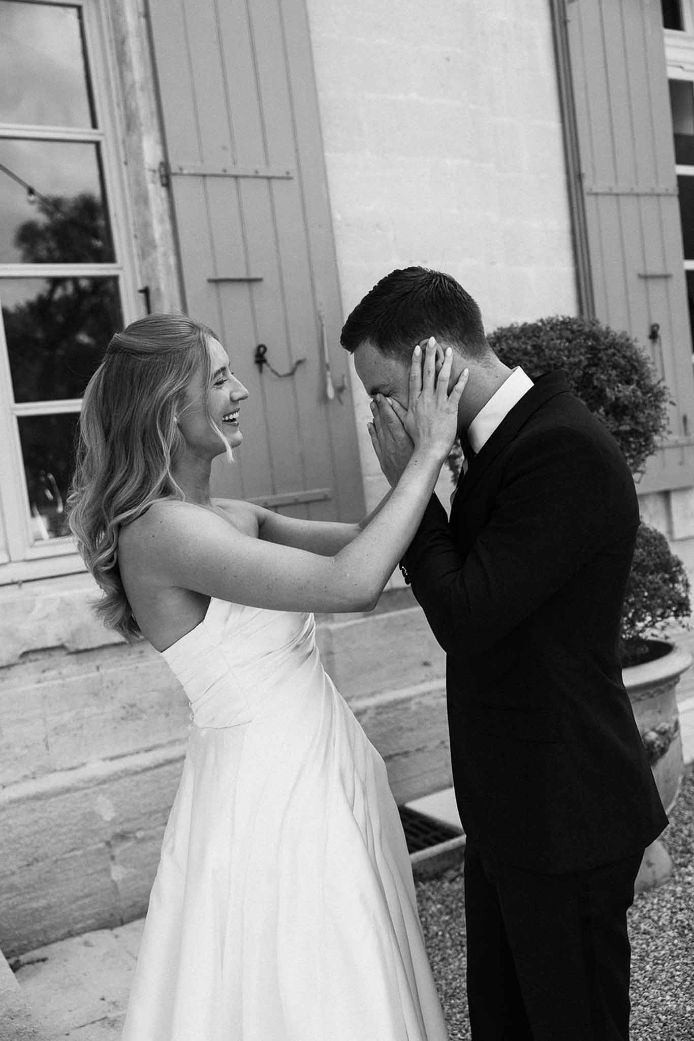 Emotional first look with groom covering face and bride laughing at chateau courtyard in black and white